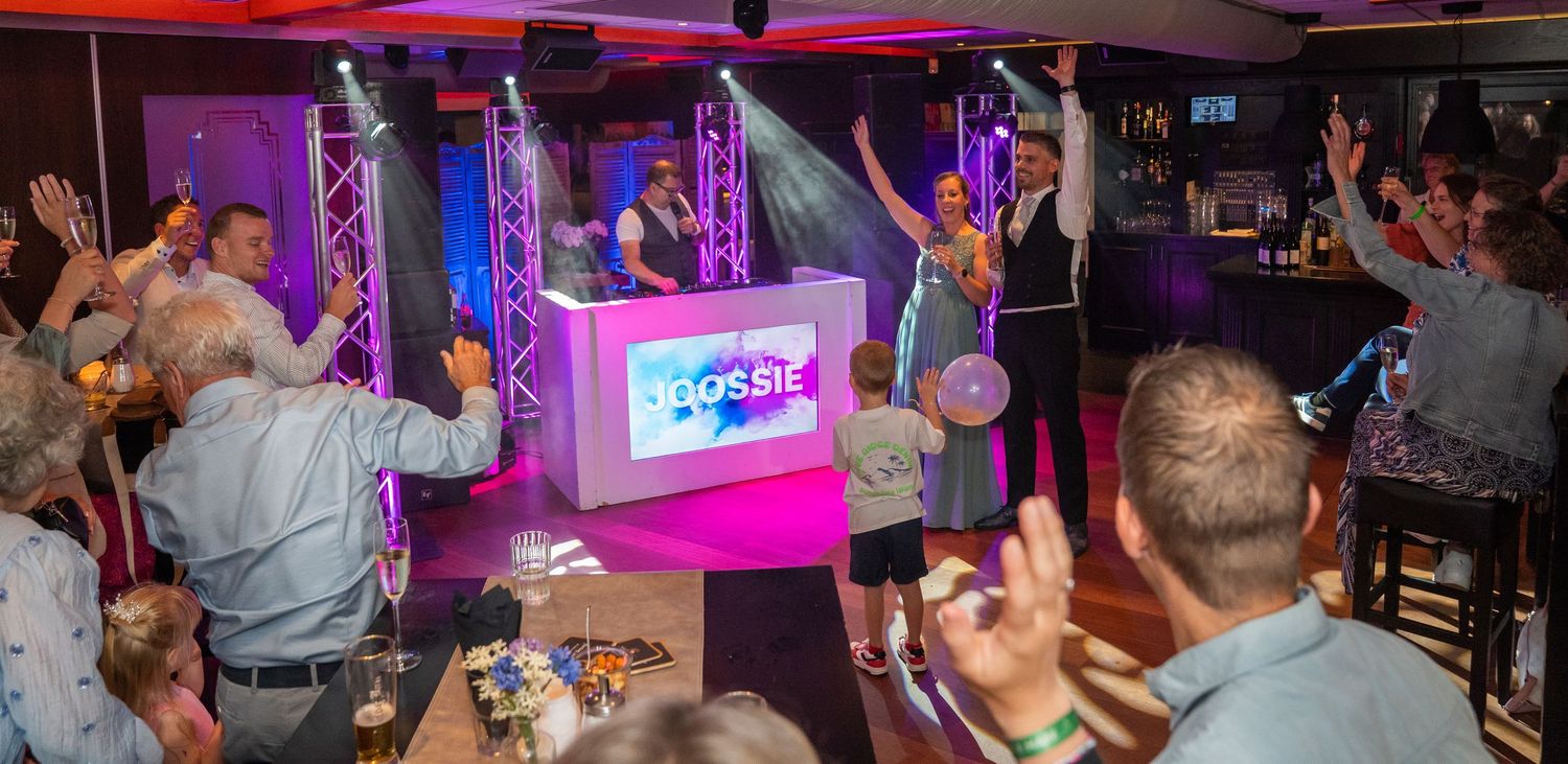 Een dj draait muziek op het podium terwijl een enthousiast publiek danst en de handen in de lucht steekt tijdens een levendig indoorfeest.
