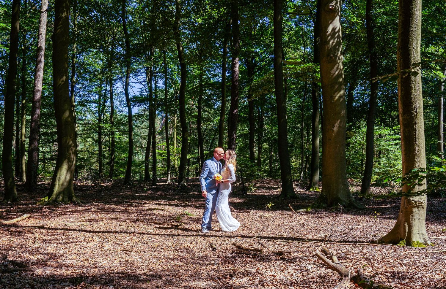 Bruid en bruidegom kussen elkaar in een zonovergoten bos, omringd door hoge bomen en herfstbladeren op de bosbodem.