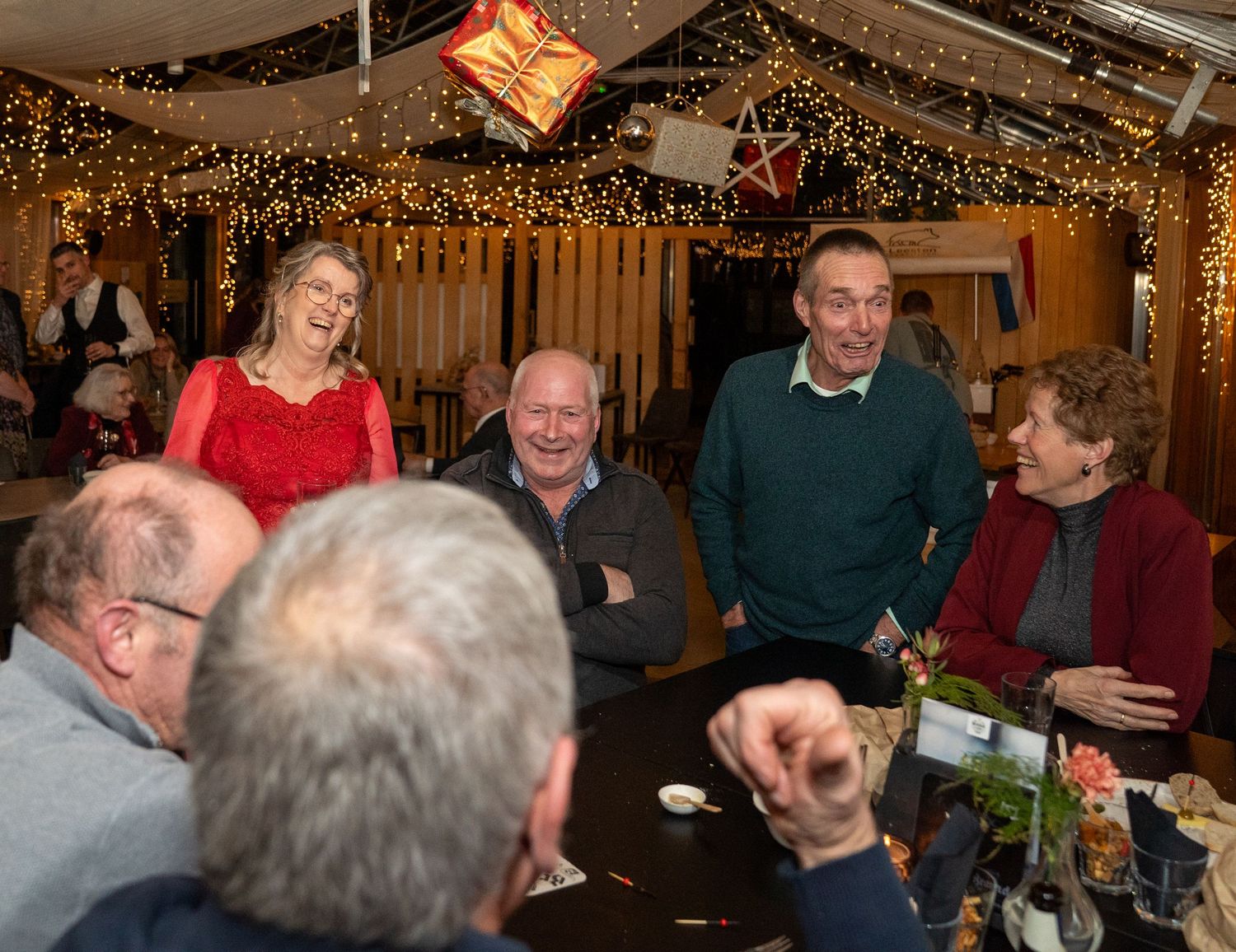 Een vrolijke groep gasten lacht en praat gezellig op een feestelijk kerstfeest met fonkelende lichtjes.