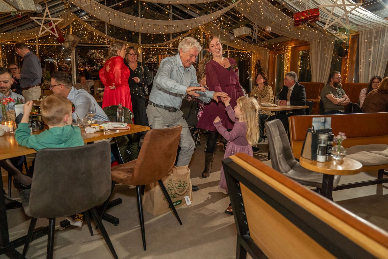 Familiebijeenkomst in een sfeervol restaurant met lichtjes; een oudere man begroet een jong kind tussen de tafels door.