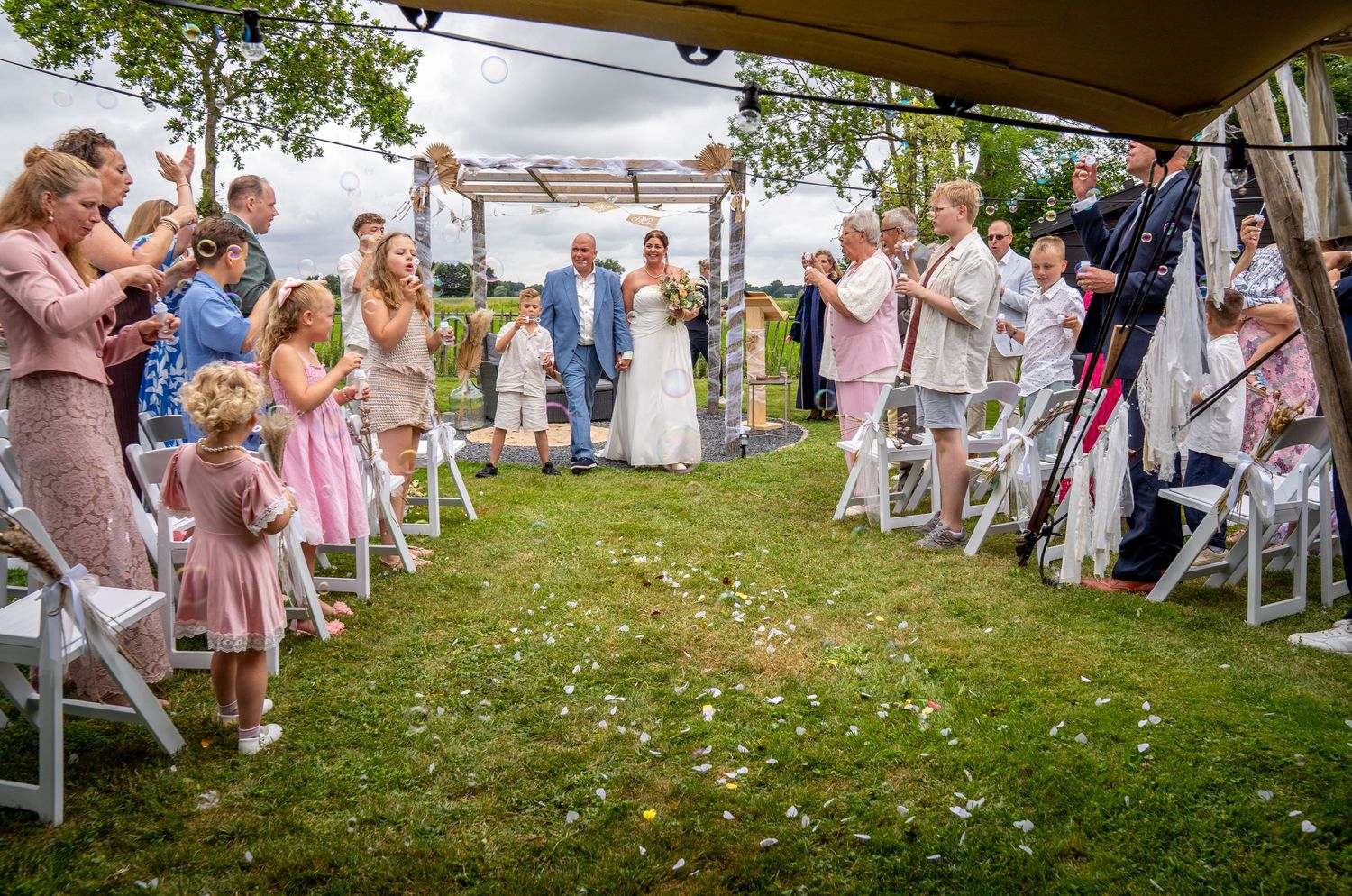 Outdoor wedding ceremony with guests tossing flower petals as couple walks down aisle on green lawn.