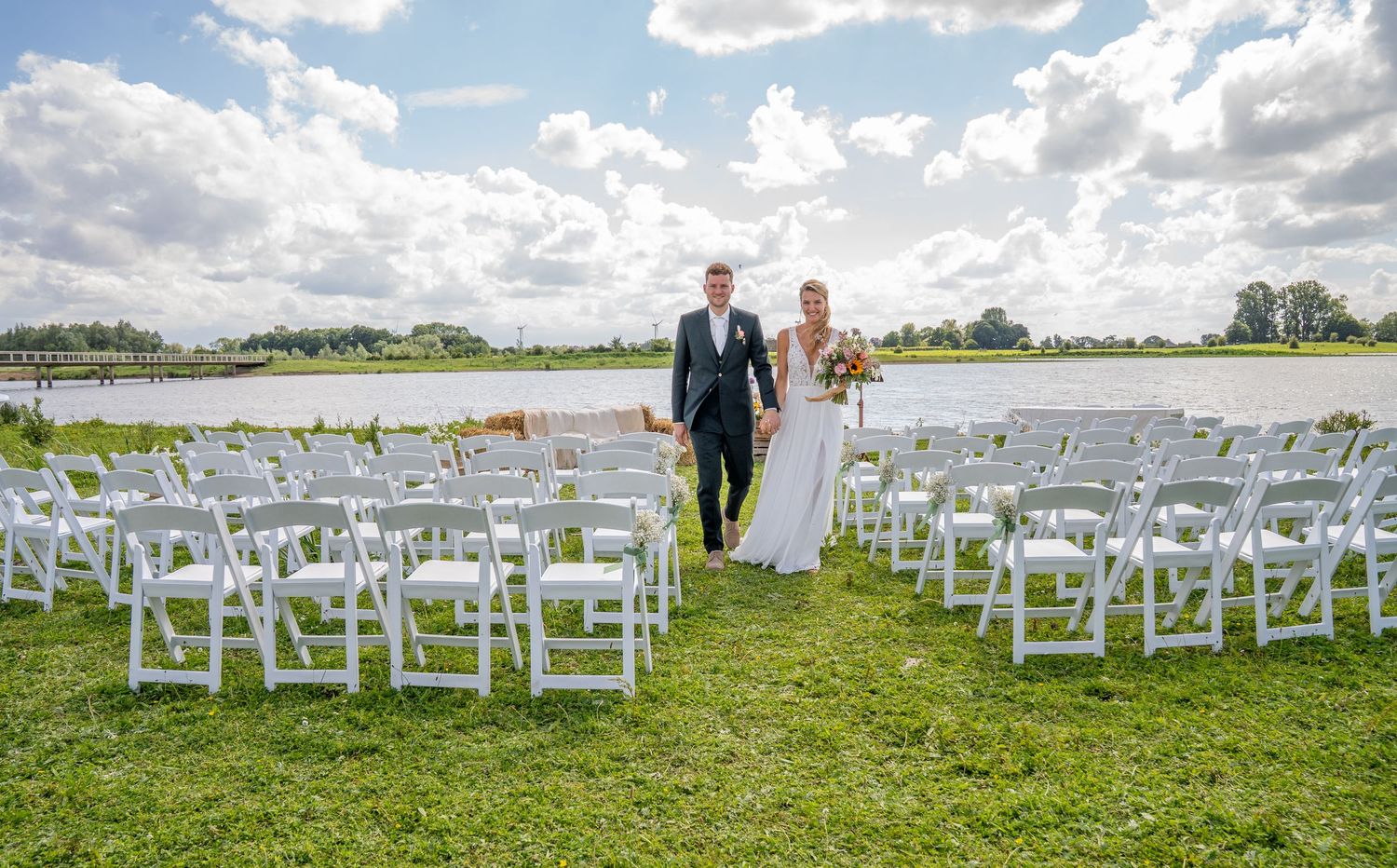 Bride and groom walk hand-in-hand down outdoor aisle lined with white chairs beside a scenic waterfront.