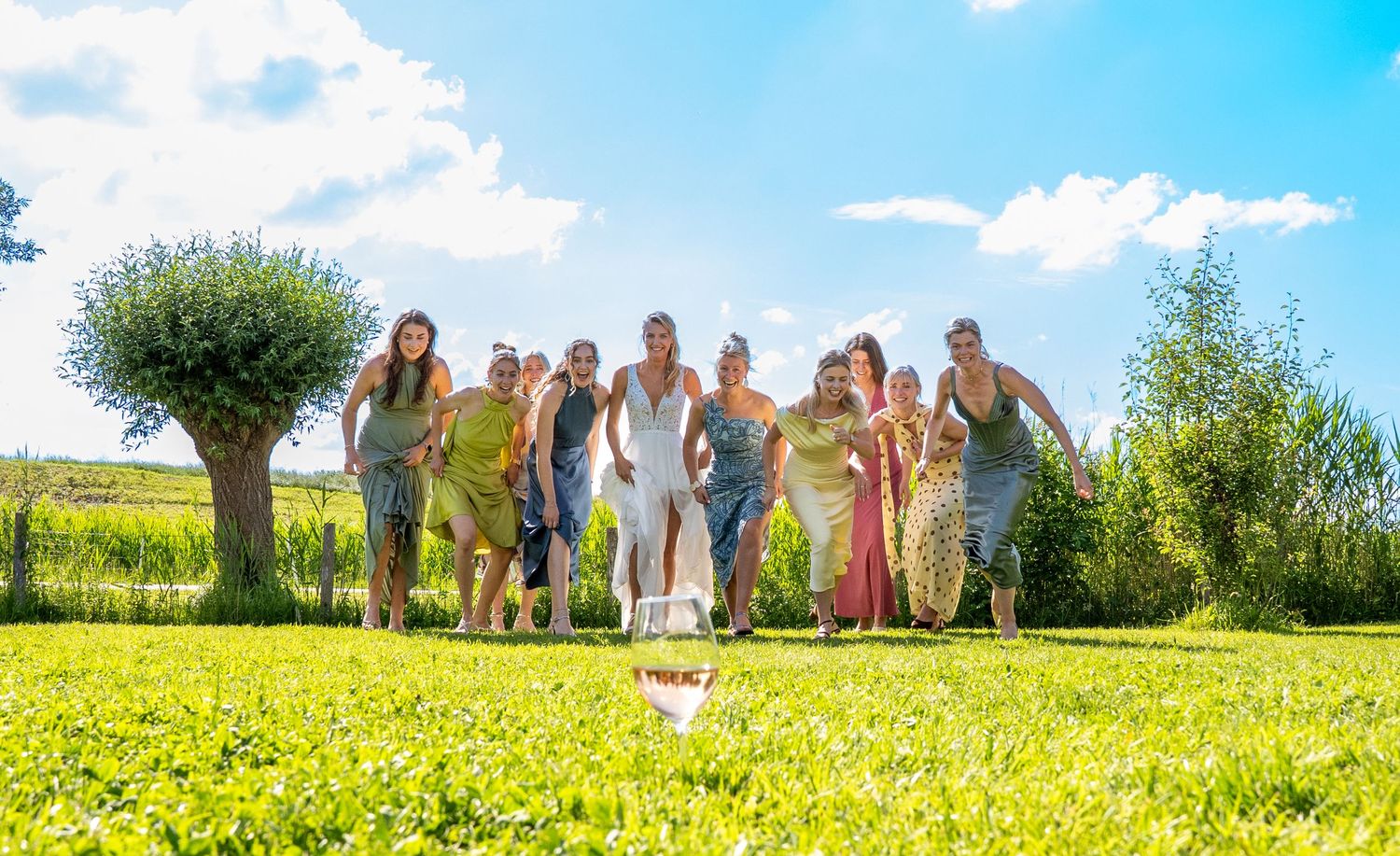 A joyful bridal party runs across a lush green lawn under a bright blue sky on a sunny wedding day.