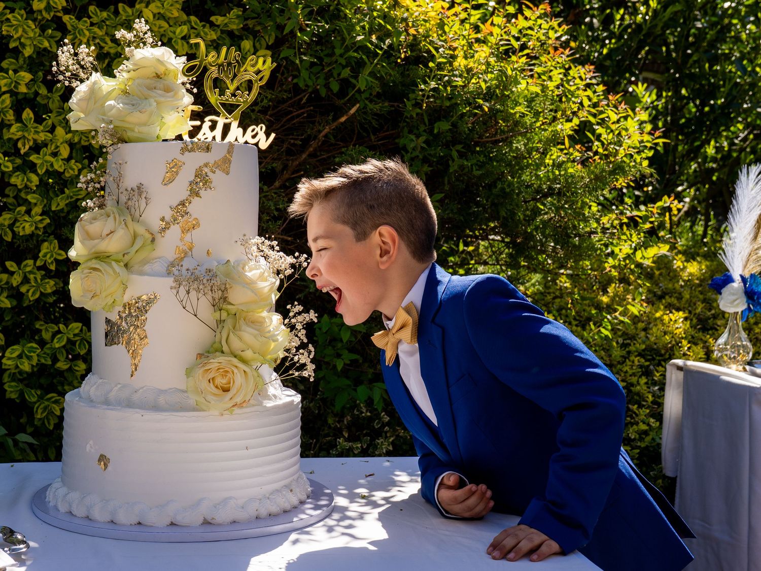 A young boy in a blue suit leans toward a tiered white wedding cake decorated with flowers outdoors.
