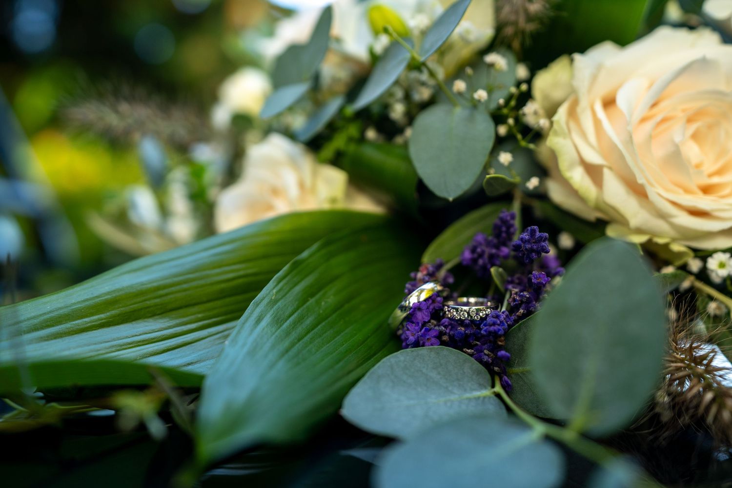 Close-up van gouden trouwringen die rusten op groene bladeren naast crèmekleurige rozen en paarse bloemen in een boeket.