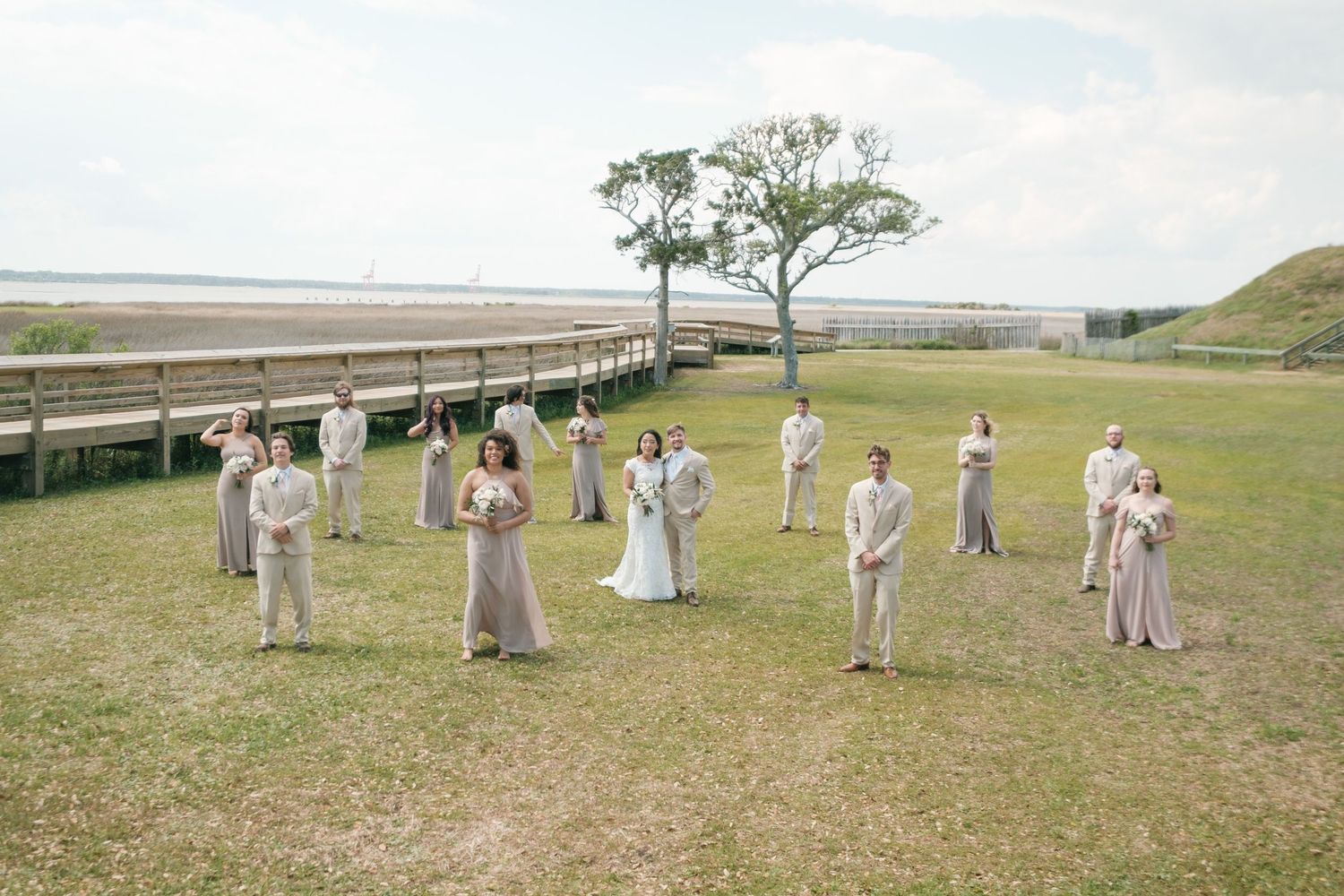 A wedding party in soft gray attire stands arranged on a grassy hilltop overlooking a scenic coastal landscape.