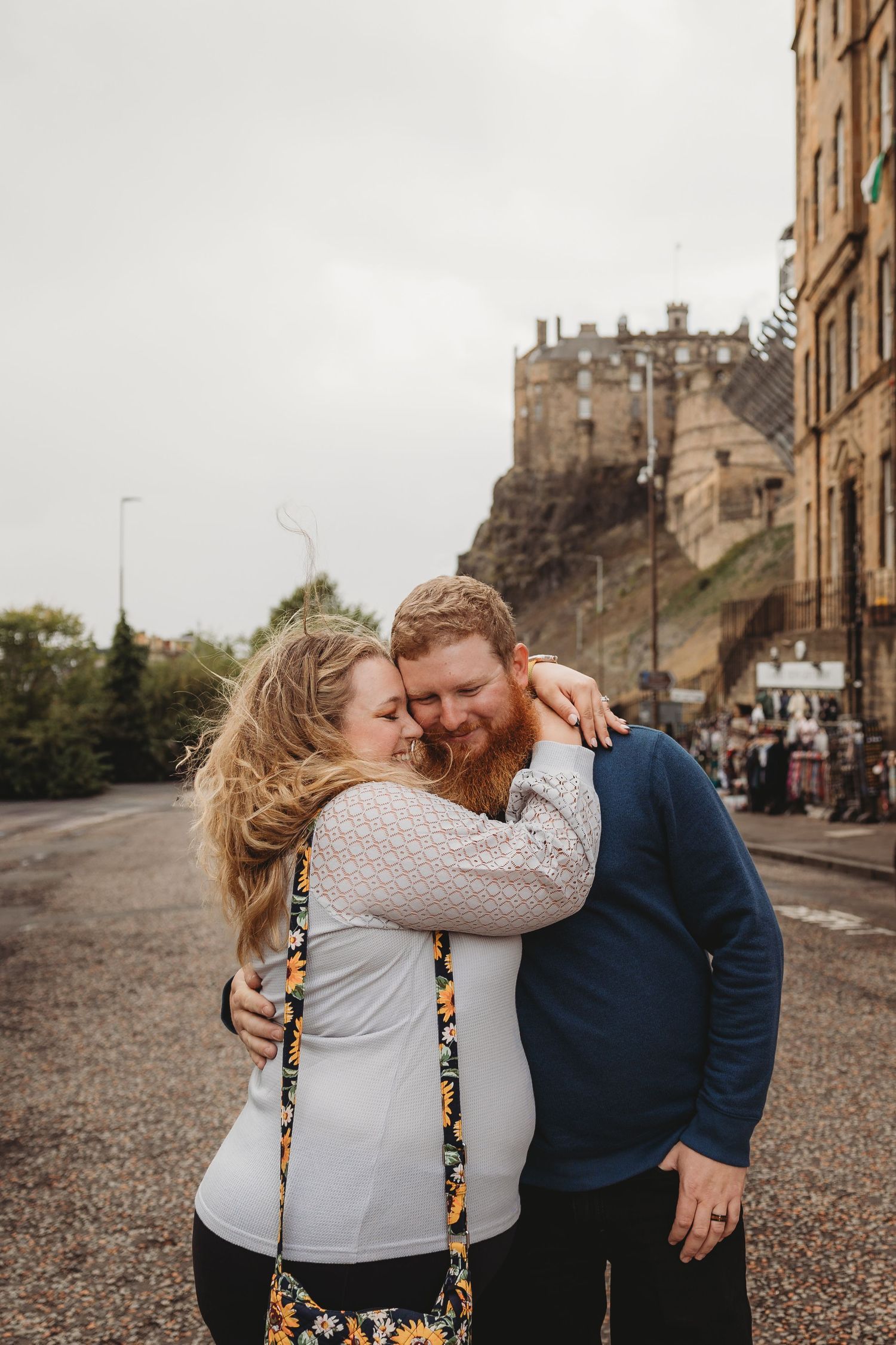 Couple hugging on a street near Edinburgh Castle, woman in white lace top, man in blue sweater, overcast sky.
