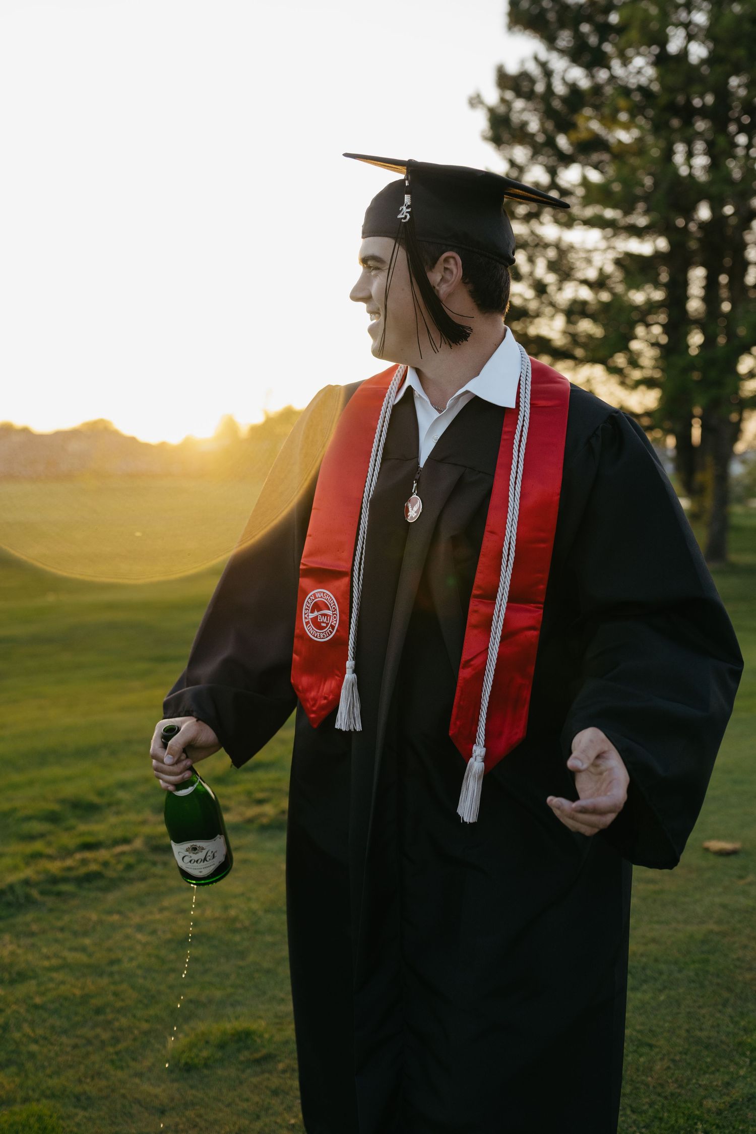 Graduate in black cap and gown with red stole pours champagne outdoors at sunset.