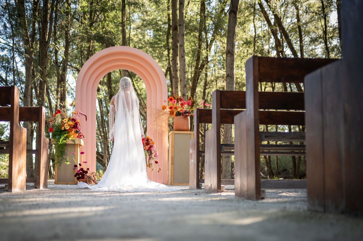 Bride standing before a pink arch in an outdoor woodland wedding ceremony with wooden pews and floral arrangements.