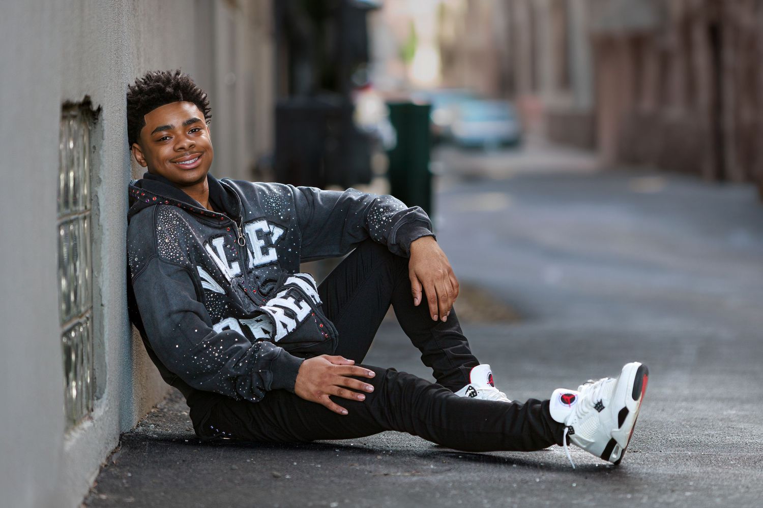 A person in a dark hoodie sits casually against a wall on an urban street wearing white sneakers.