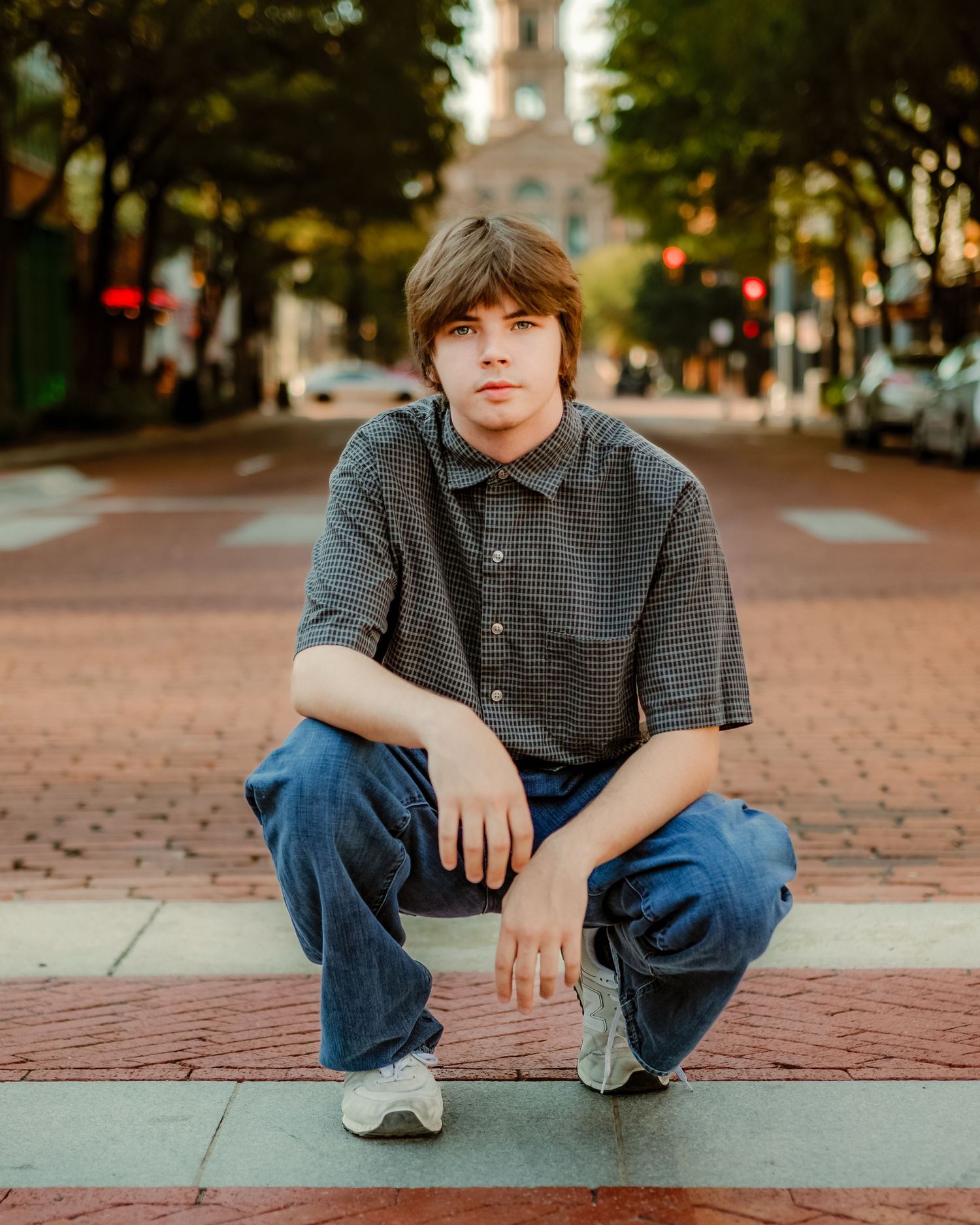 A young man in jeans and striped shirt crouches casually on brick pavement in an urban setting at sunset in Fort Worth, TX.