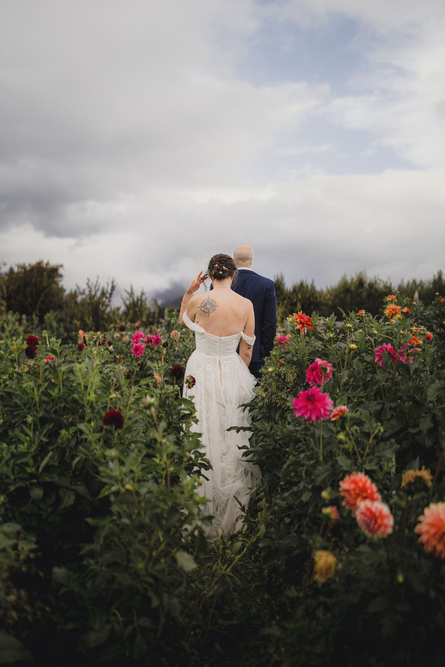 A couple in wedding attire embraces among vibrant dahlias and greenery in a garden setting under cloudy skies.