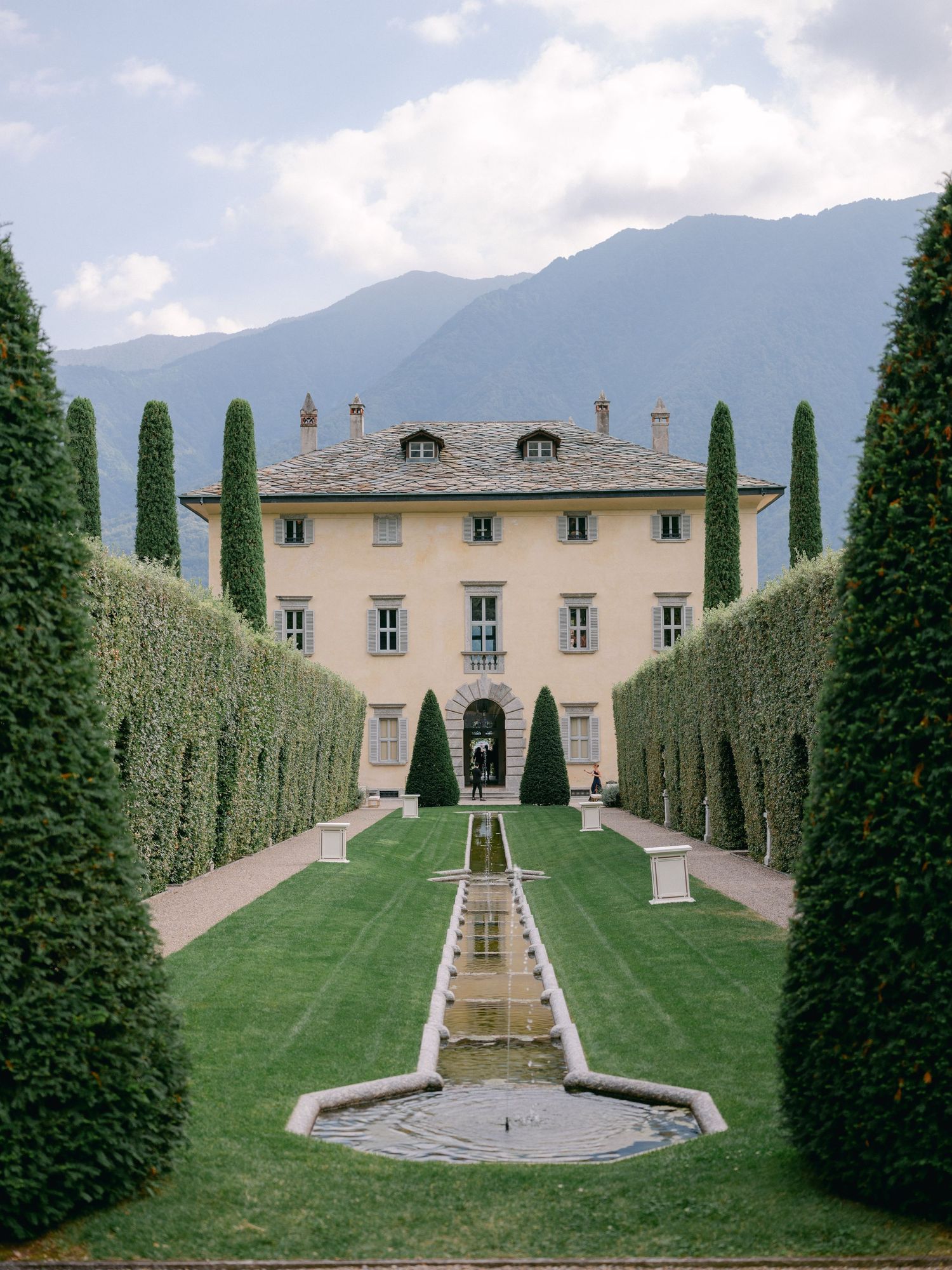 A grand historic mansion with ornate architecture and manicured gardens hosts an elegant outdoor gathering of people.