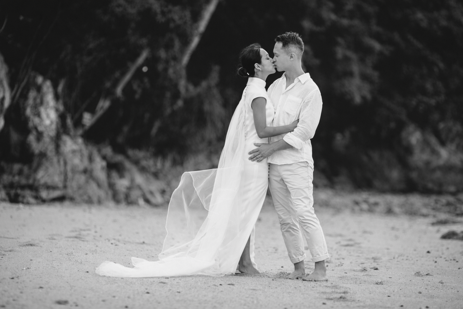 Black and white pre-wedding portrait of couple kissing on a quiet beach, soft movement in dress, intimate and timeless destination imagery.