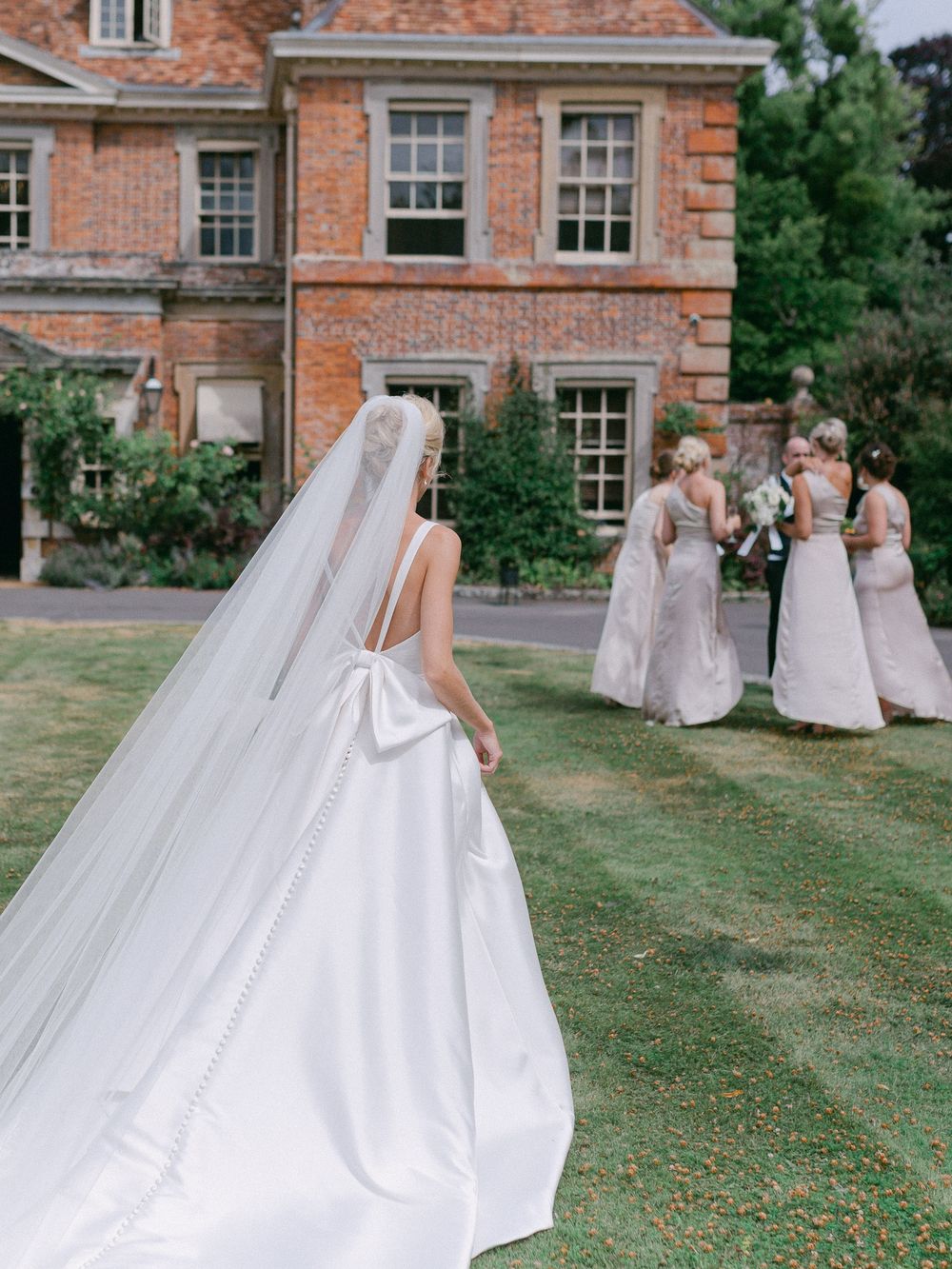 A bride in a flowing white dress with bow detail stands on the lawn of a brick mansion.