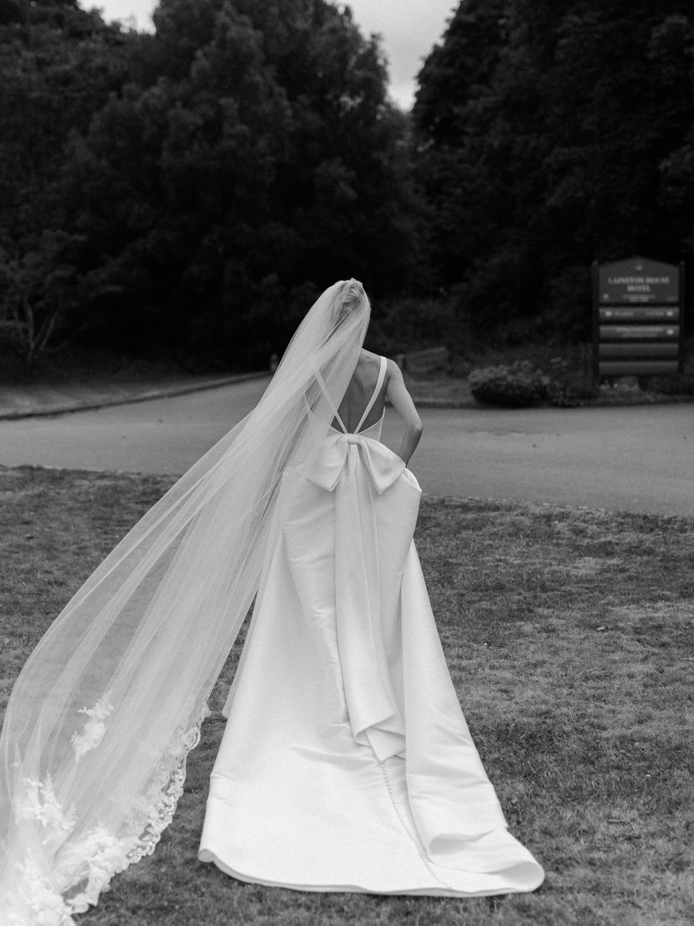 A bride in an elegant white gown with long veil stands on a path surrounded by trees.
