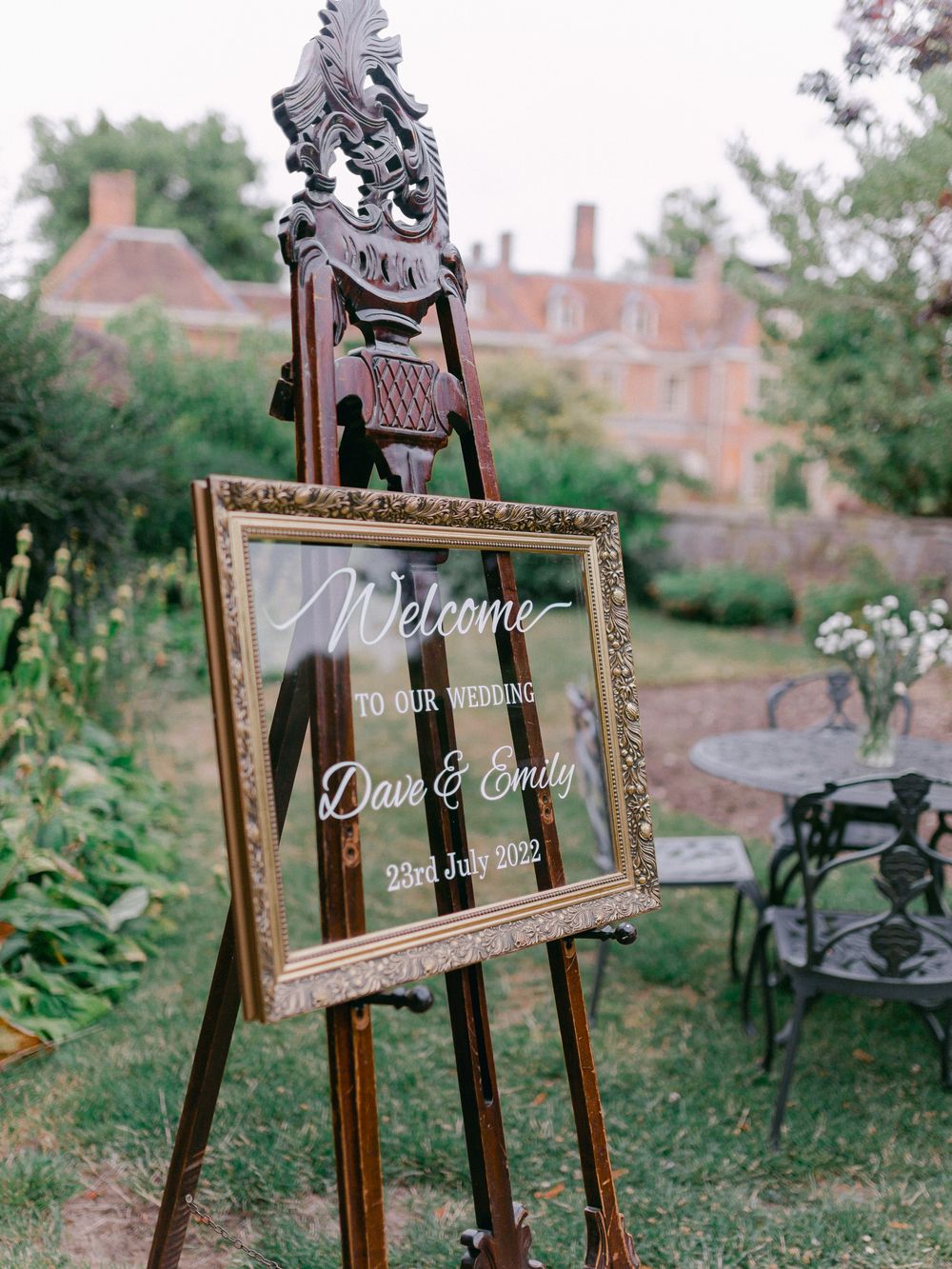 An ornate welcome sign on an easel in a garden setting with a mansion in the background.