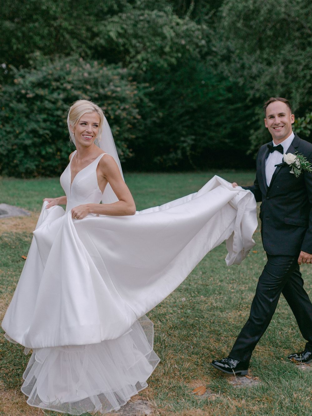 A bride in a flowing white dress twirls playfully on a grassy lawn while the groom watches in a classic black tuxedo.