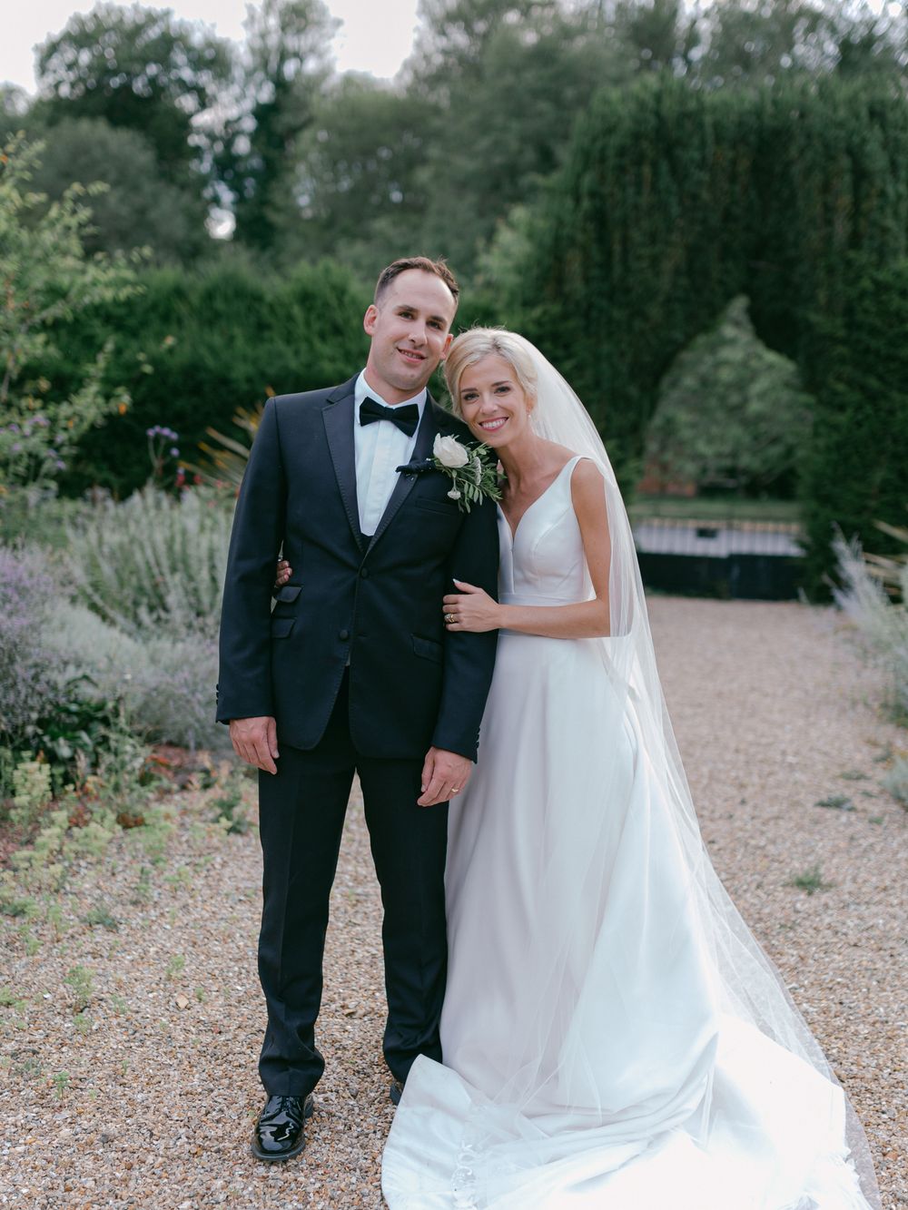 A wedding couple poses on a gravel path surrounded by manicured garden landscaping.