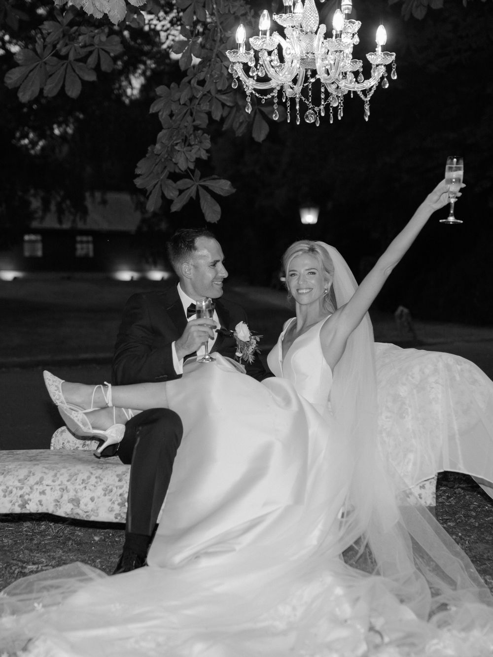 A couple celebrates at their wedding reception under a crystal chandelier, with the bride raising her champagne glass in joy.