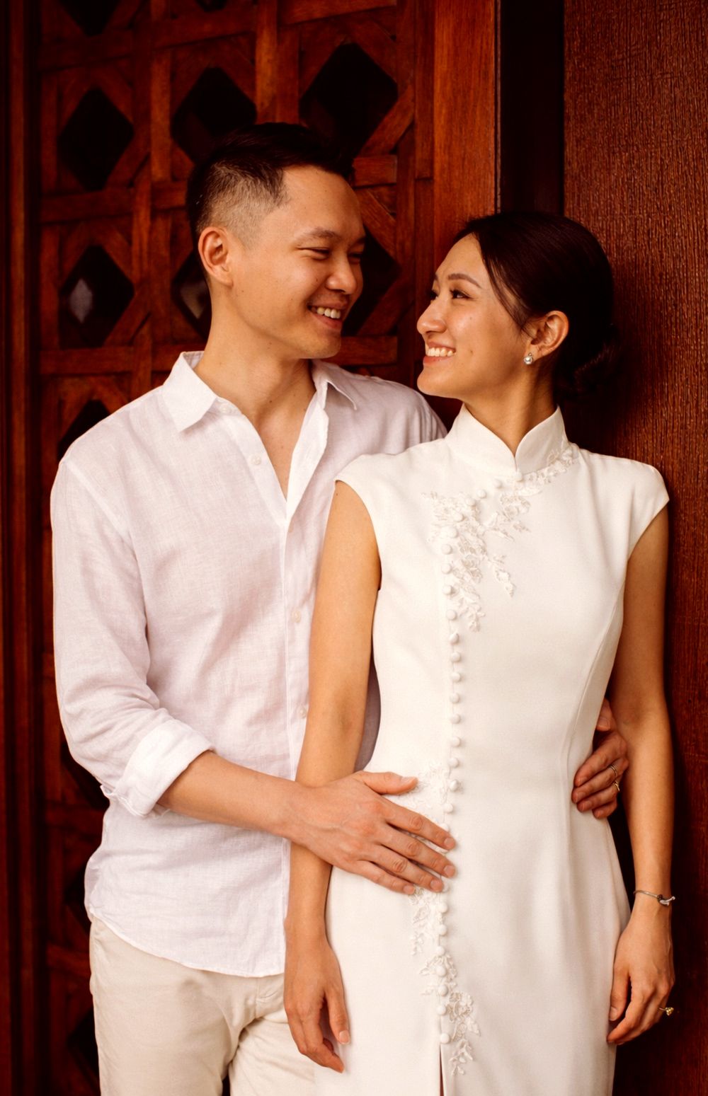 Couple in traditional attire at Thai villa doorway, smiling and embracing, warm light, refined pre-wedding portrait with architectural depth
