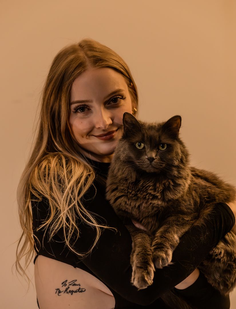 Young woman with long blonde hair smiles holding a fluffy dark cat with green eyes against a beige background.