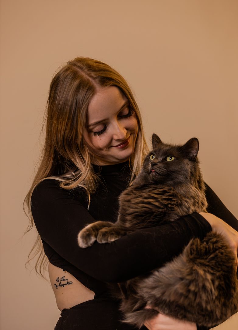 Smiling young blonde woman in black top cuddles a large fluffy dark cat with green eyes against beige backdrop.
