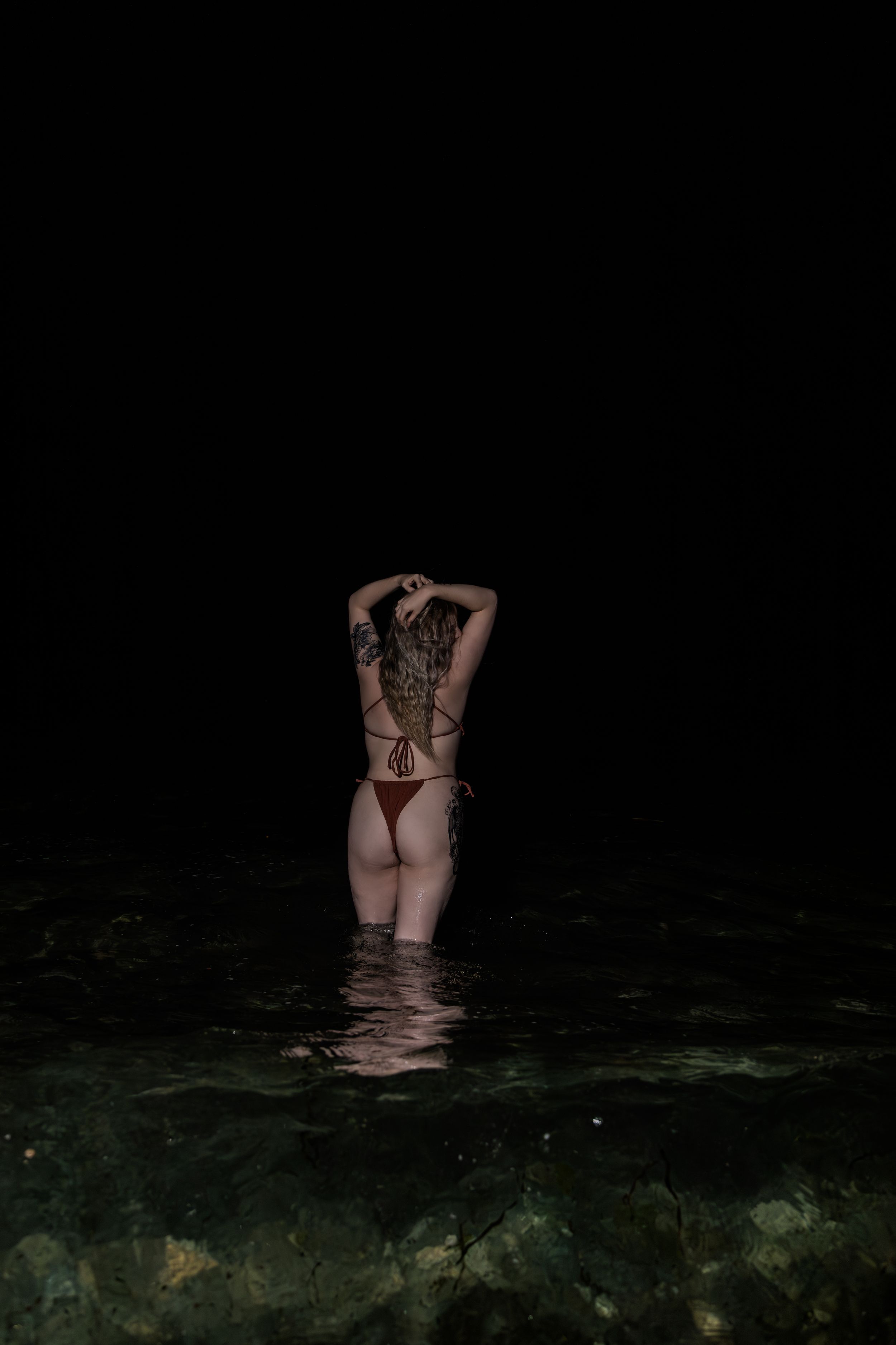 Woman with back tattoo in red bikini stands in shallow moonlit water at night facing away from camera.