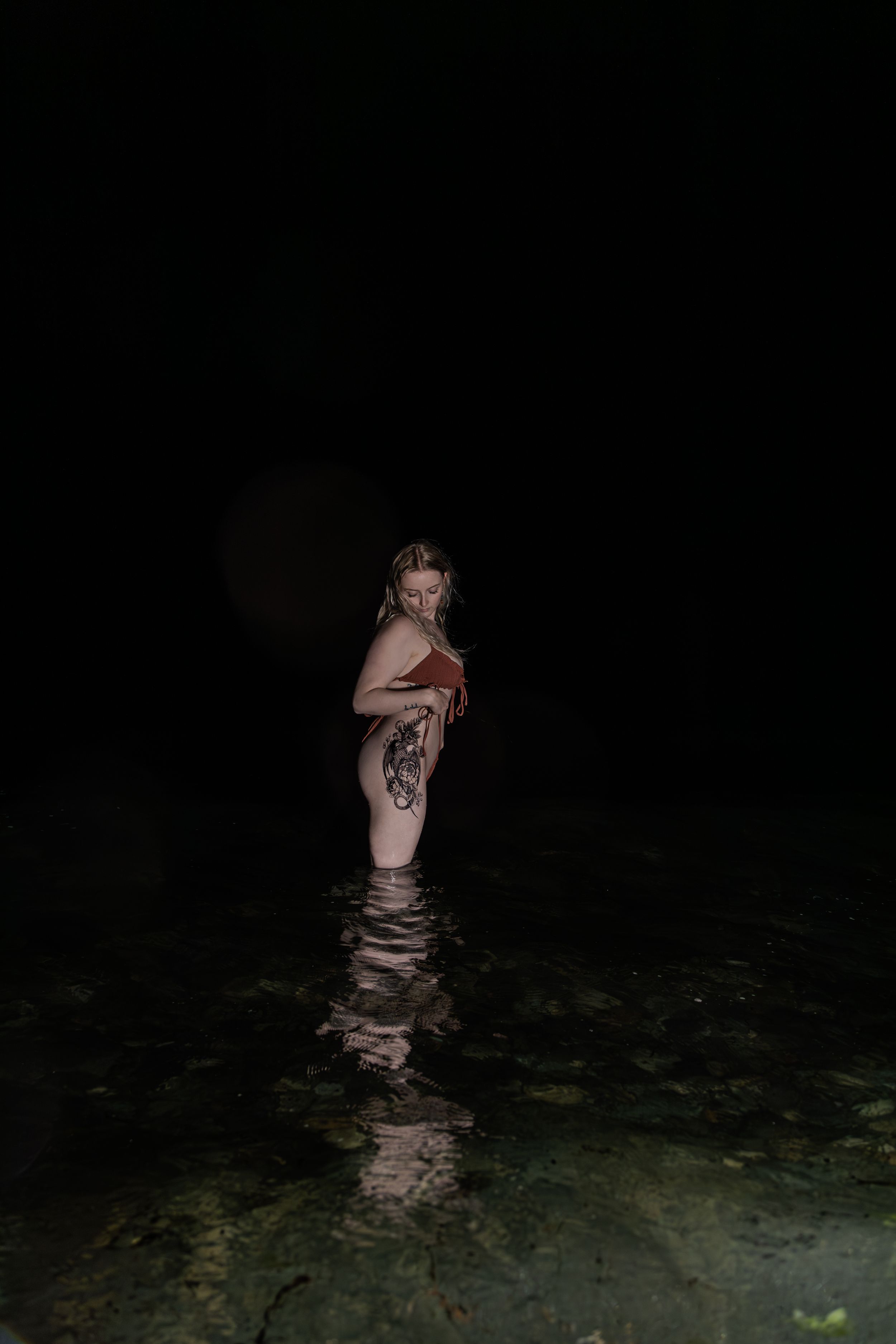Tattooed woman in red bikini stands in shallow water at night with large tattoo visible on her side.