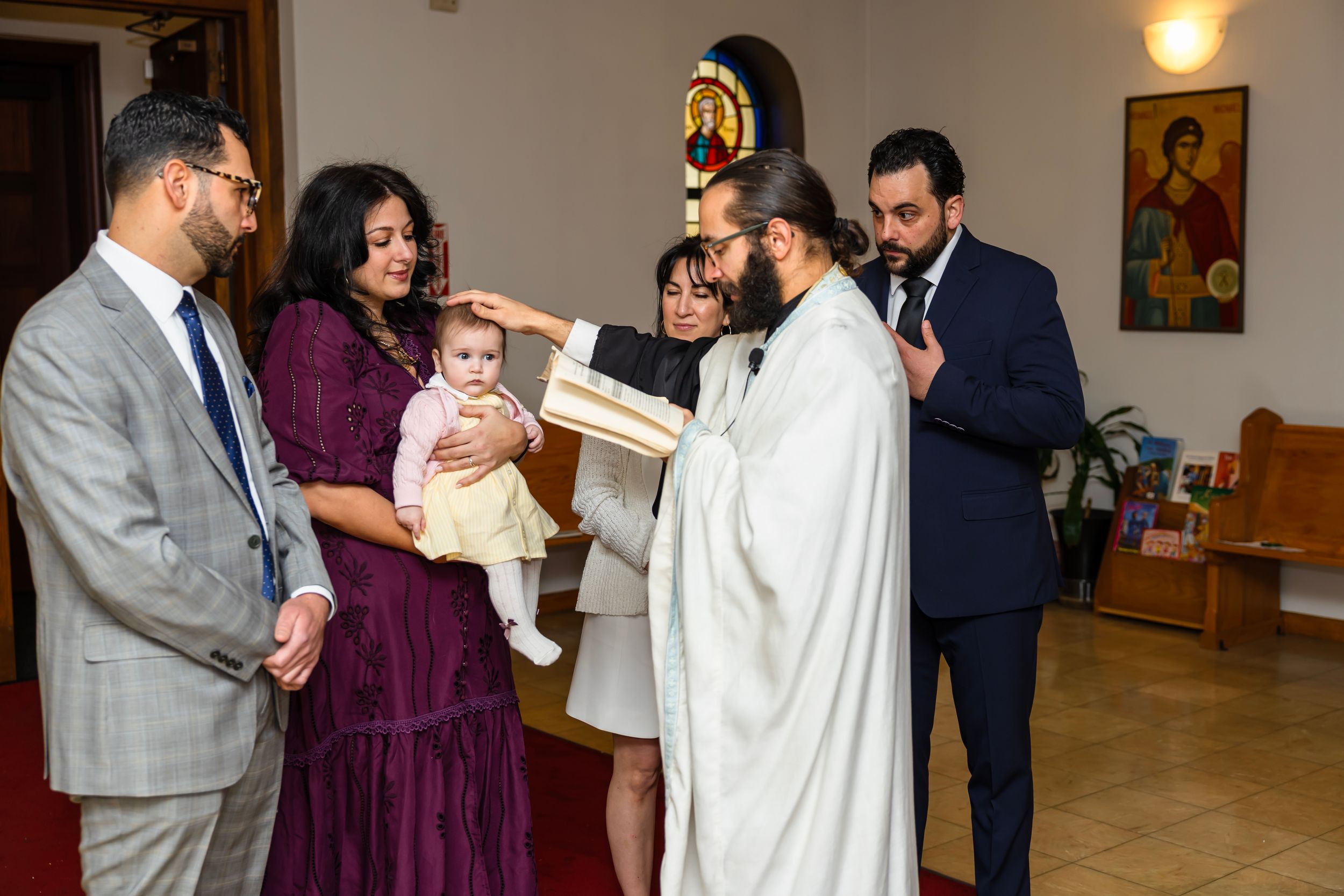 A priest performs a baptism ceremony on an infant held by parents in a church setting.