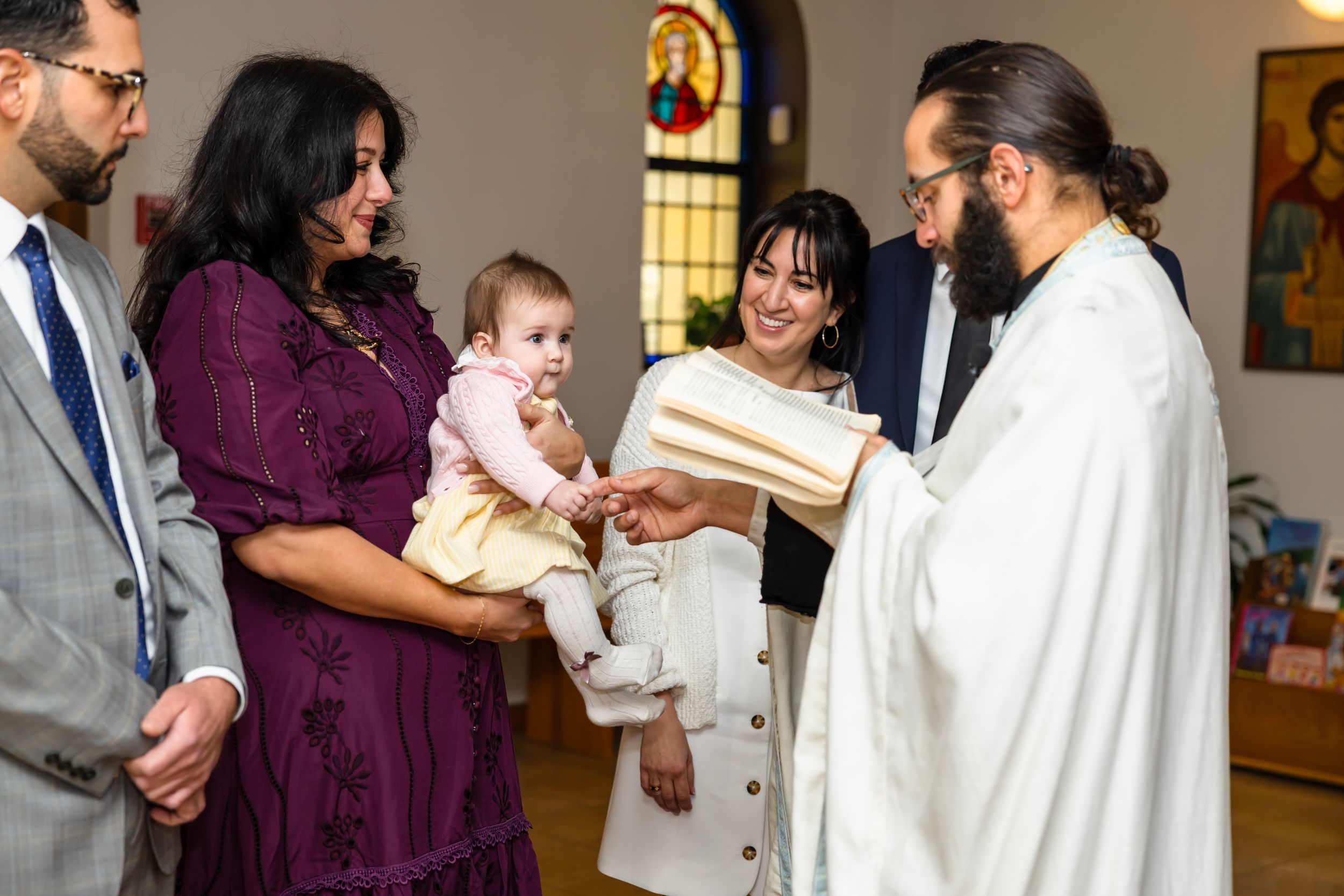 Priest in white robes performs a baptism ceremony, blessing an infant held by a woman in a purple dress.