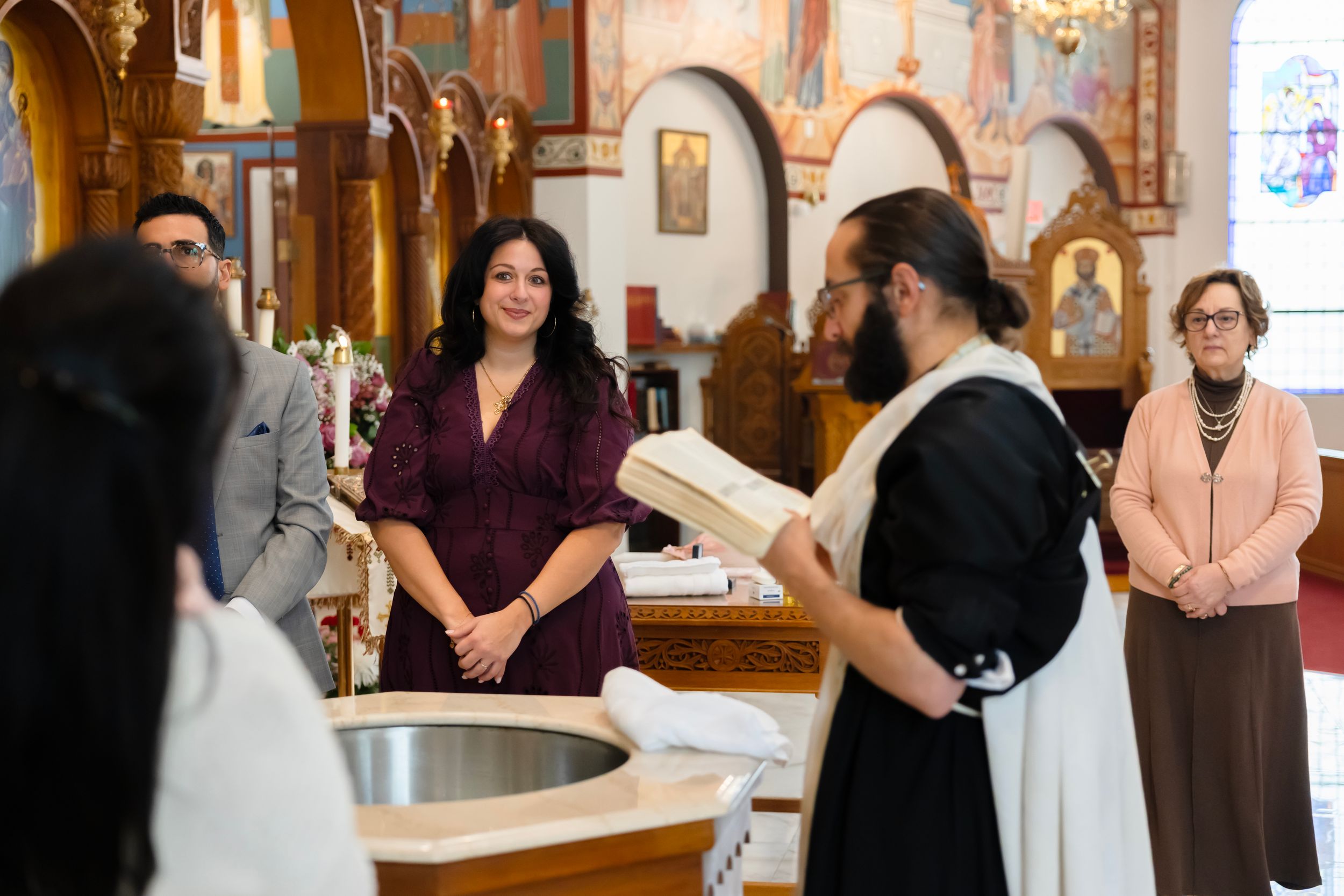 Orthodox priest reads from holy book during baptism ceremony in ornate church with attendees in formal attire.