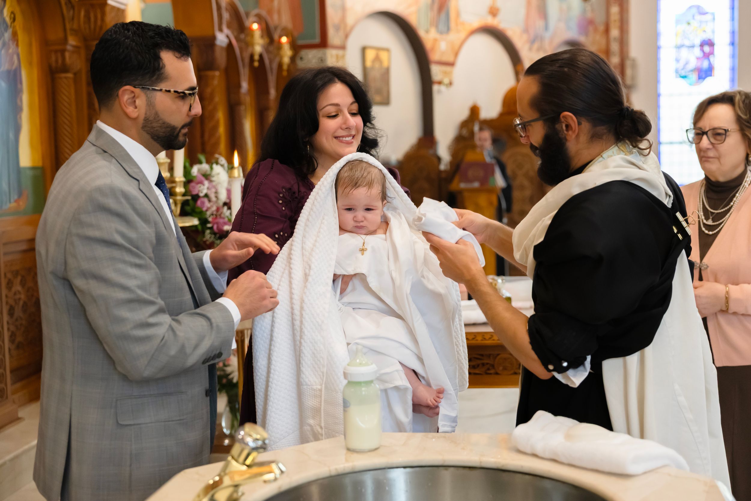 Orthodox priest baptizes baby in white gown at church font with parents watching.