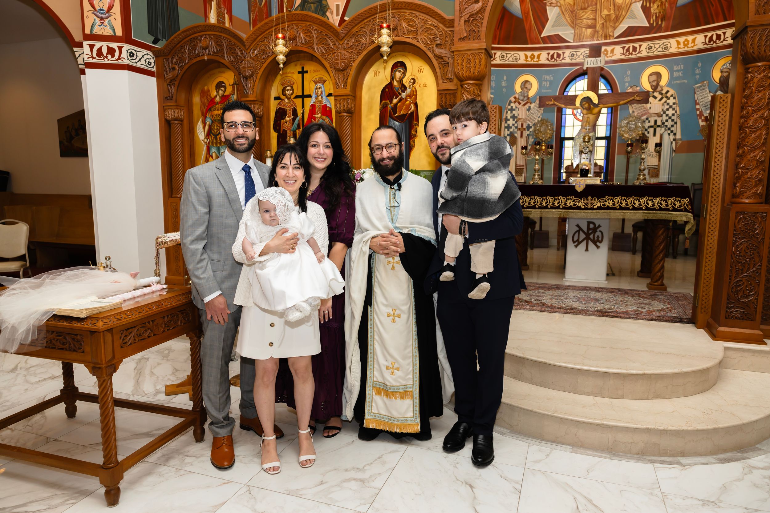 Family poses with Orthodox priest after baptism ceremony in decorated church.