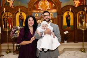 Family posing with baby in white christening gown inside an Orthodox church with golden iconostasis.