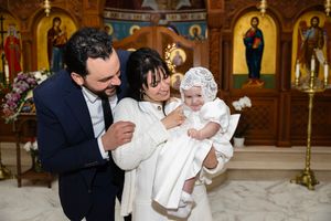 Parents proudly hold their baby dressed in white baptismal gown inside a Greek Orthodox church.