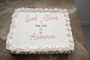 White sheet cake with pink frosting decorations, a cross, and “God Bless Arianna” written in pink icing.