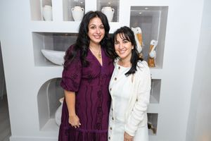 Two women smiling together, one in a purple dress and one in a white blazer, posing in front of white shelving decor.