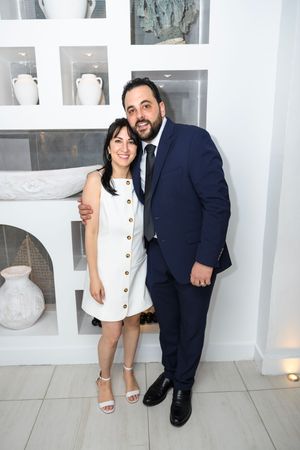 Couple posing together at elegant event, woman in white dress and man in navy suit, smiling in front of white shelving unit.