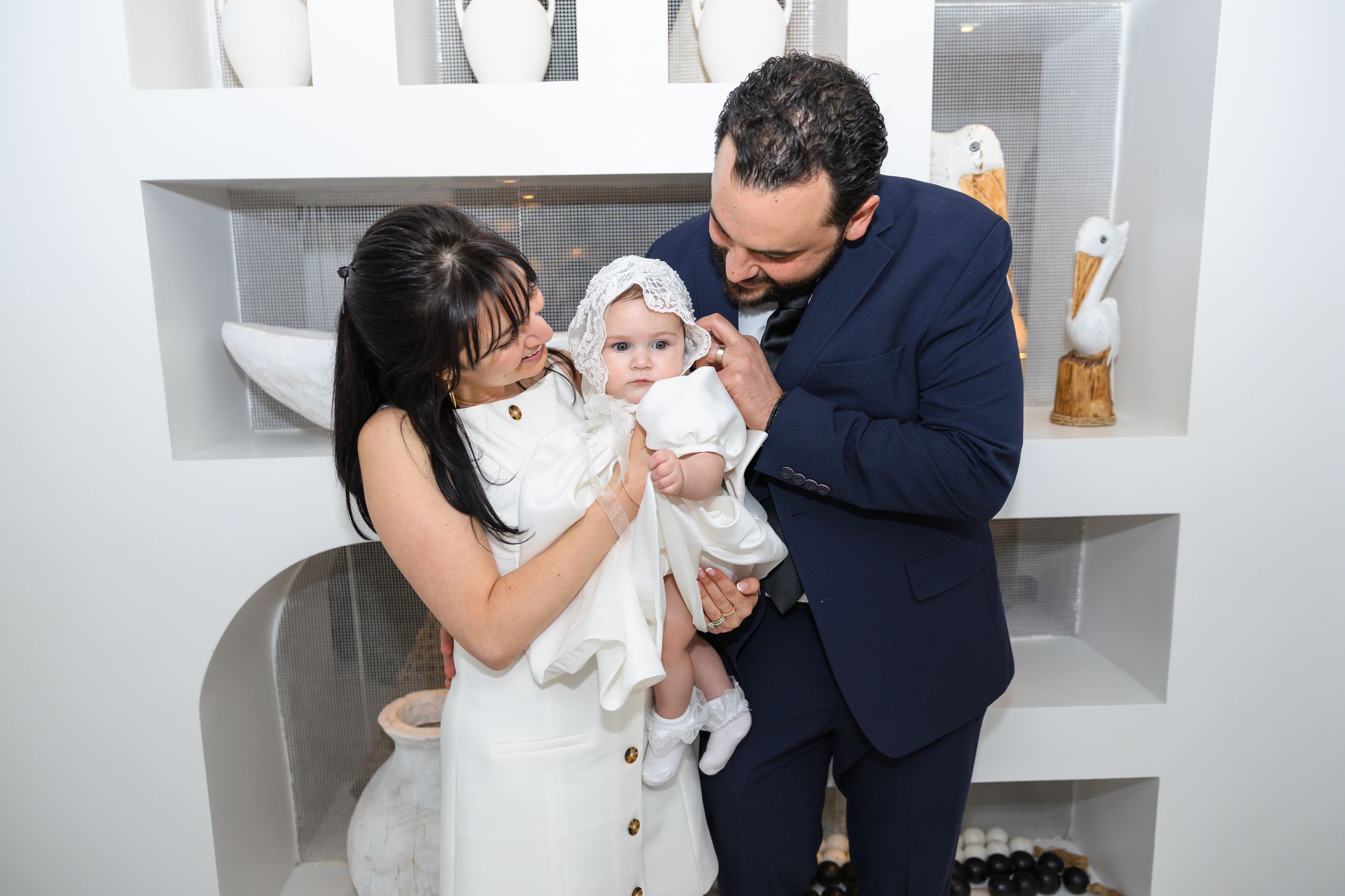 Loving parents admire their baby in white christening gown and bonnet at baptism celebration.