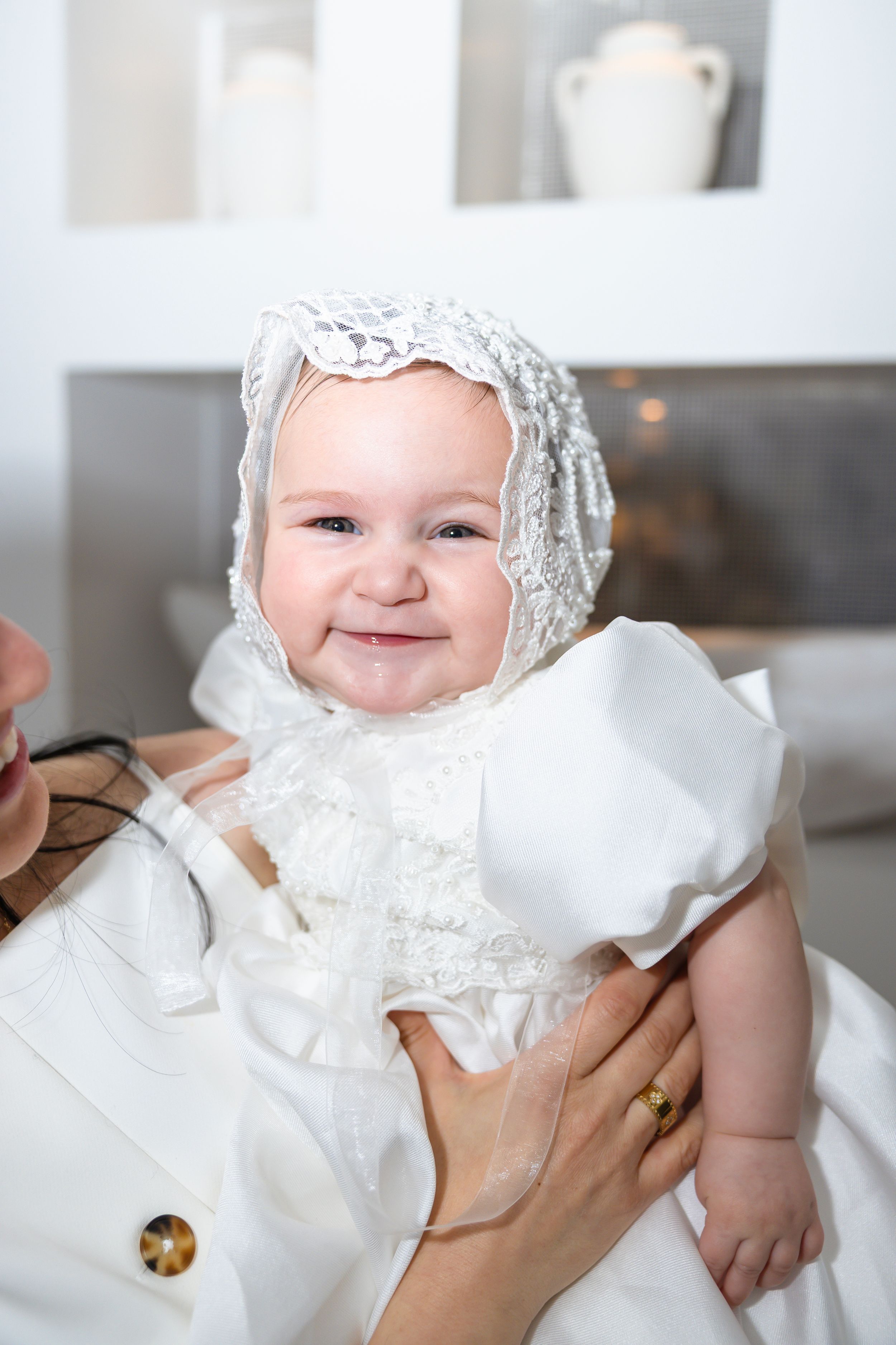 Adorable smiling baby in white christening gown and lace bonnet being held during baptism ceremony.