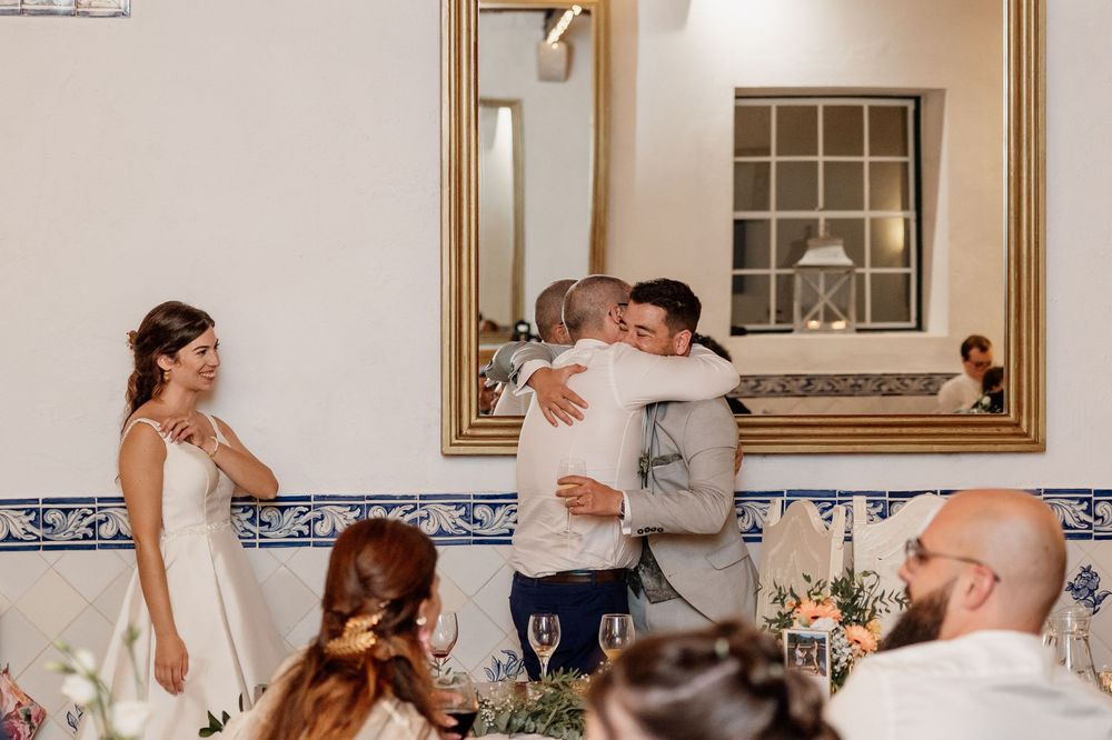 Groom hugging a guest at a wedding reception, bride smiling nearby, blue tile wall and mirror in background.