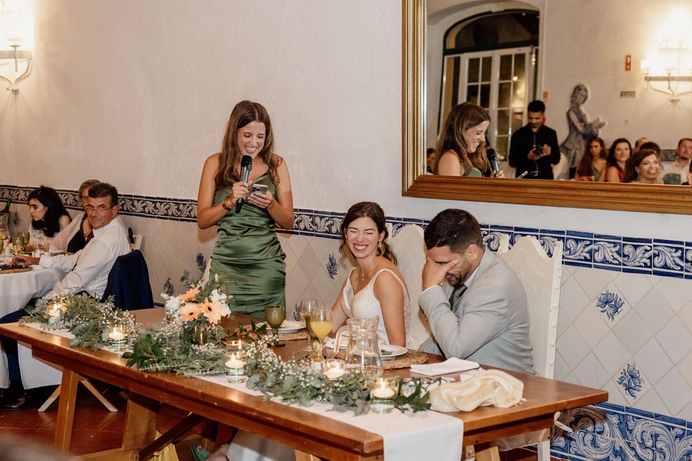 Wedding reception toast with bridesmaid speaking, bride and groom seated at candlelit table with floral centerpieces.