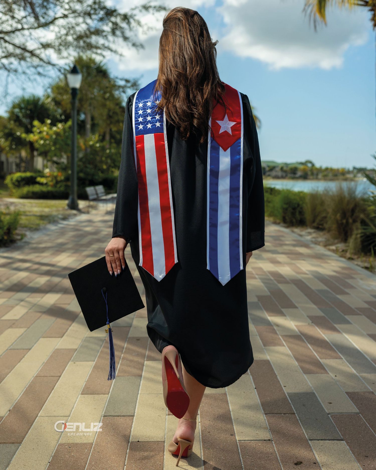Graduate in black cap and gown wearing USA and Cuban flag stole, walking on brick path outdoors.
