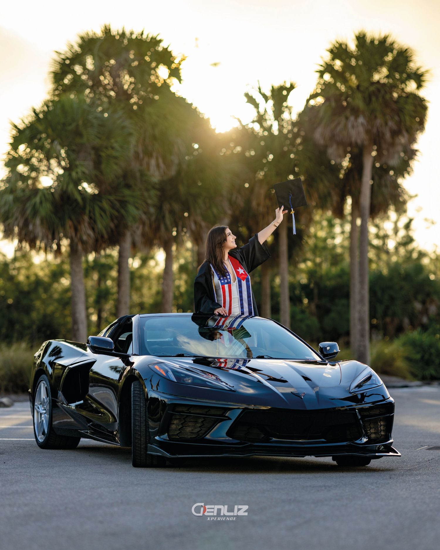 A graduate in cap and gown poses triumphantly on a black Corvette C8 at sunset with palm trees in the background.