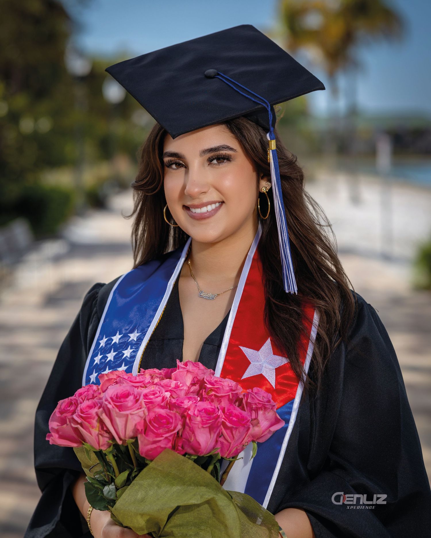 Graduate in cap and gown holding pink roses, wearing Cuban flag sash, smiling outdoors near palm trees.