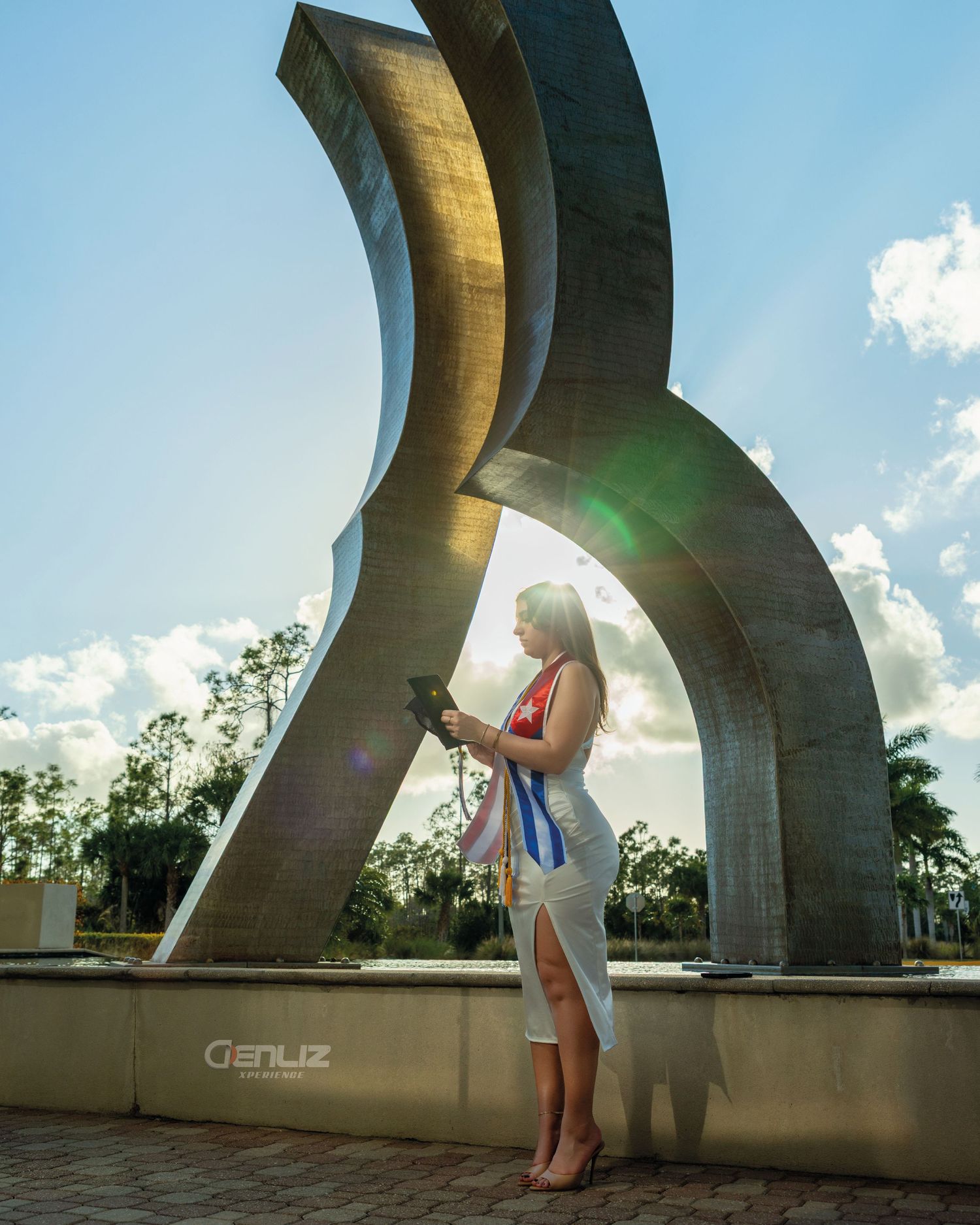 Woman in white dress stands before large abstract metal sculpture outdoors, with sun flare and blue sky backdrop.