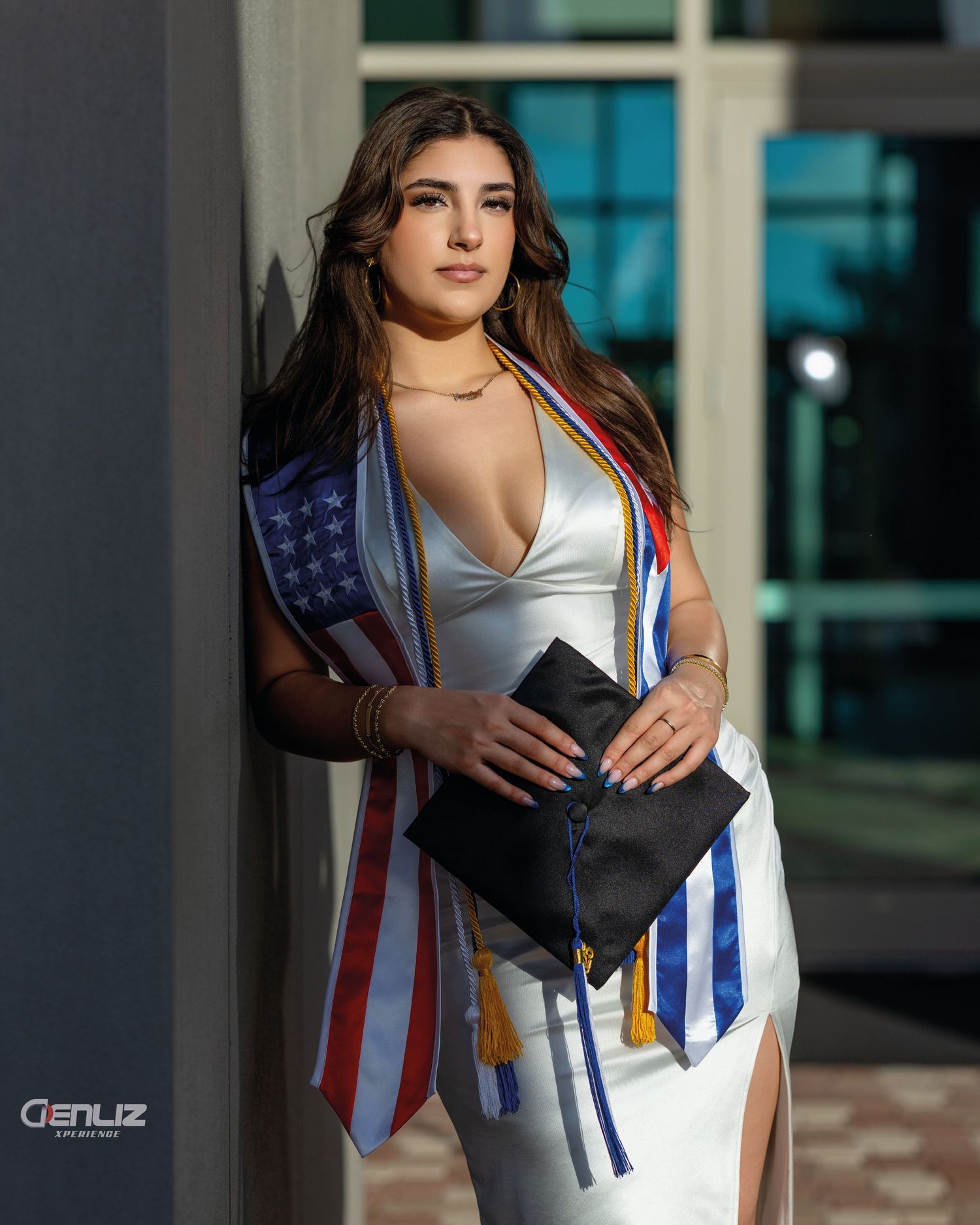 Young woman in white dress with graduation stole, American flag sash, holding diploma at outdoor ceremony.