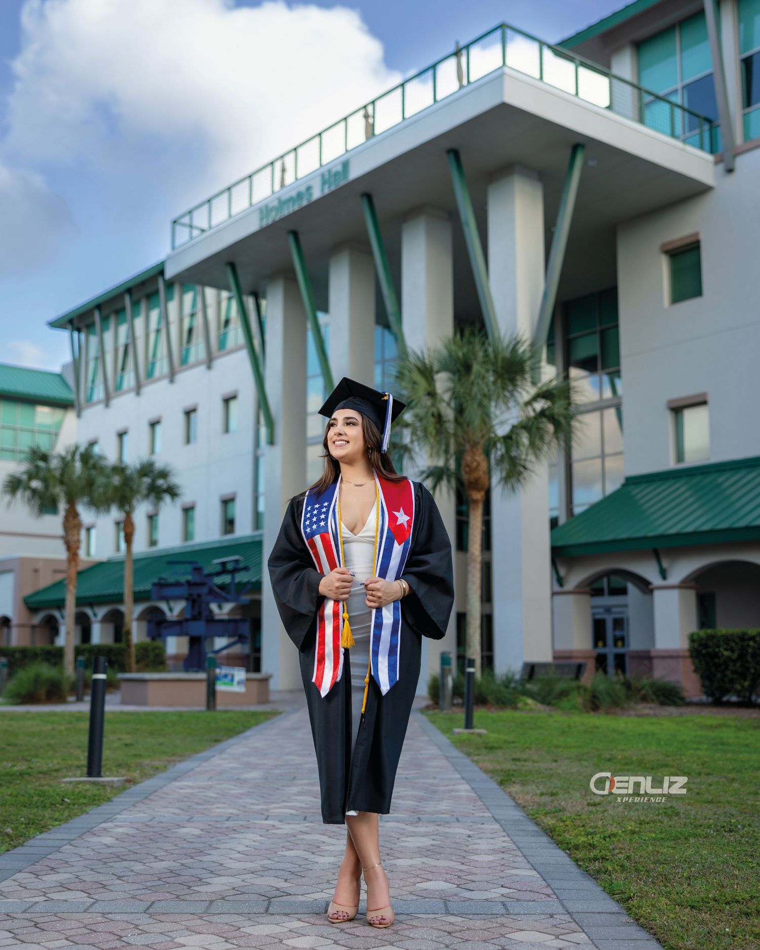 A smiling graduate in black cap and gown with colorful stoles stands on a campus walkway with a modern building behind her.