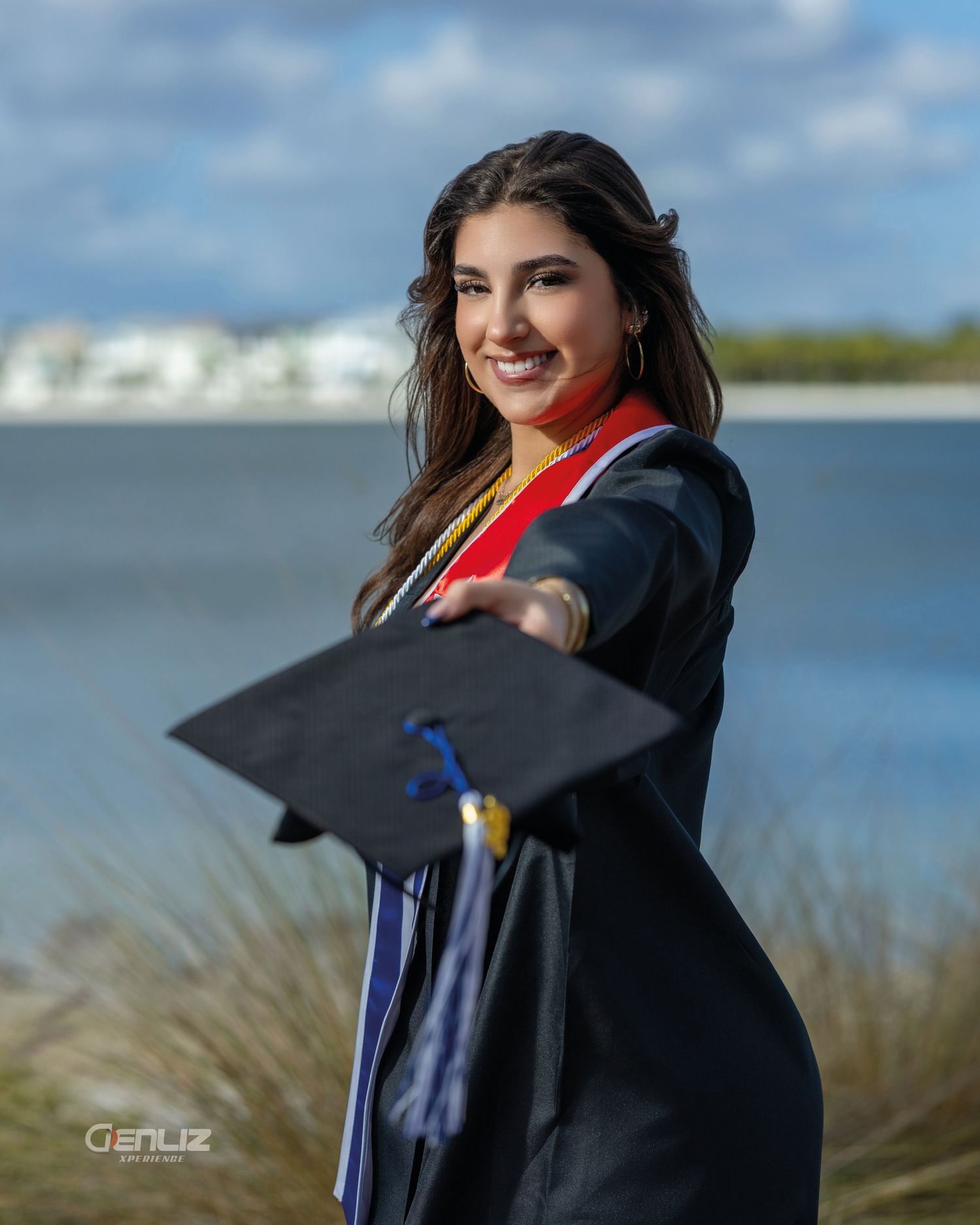 Smiling female graduate in black cap and gown holding mortarboard toward camera outdoors near waterfront.